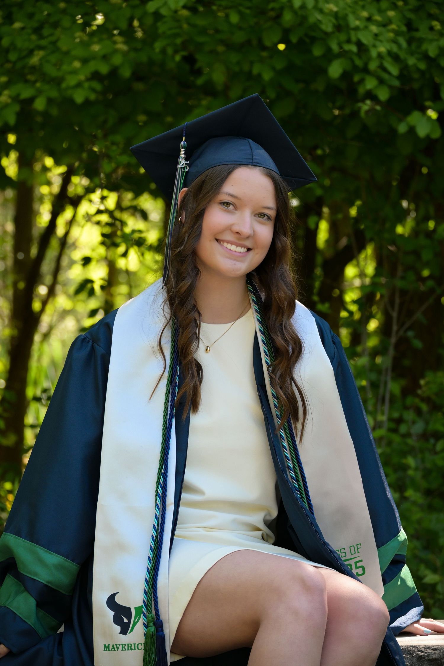 Graduate in dark academic gown and white stole sits outdoors for portrait session.