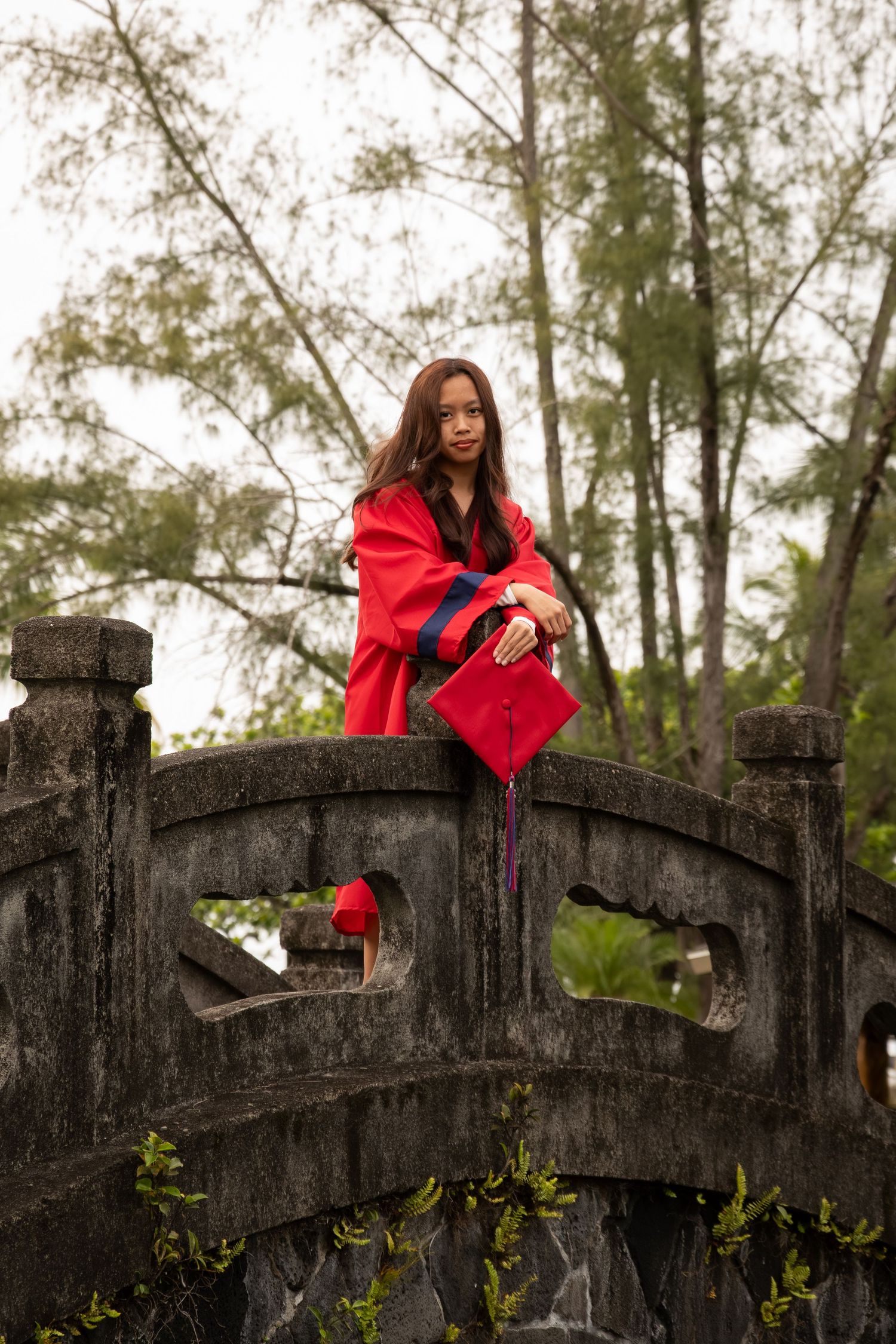 Young woman in red graduation gown and cap posing on ancient stone bridge surrounded by trees.