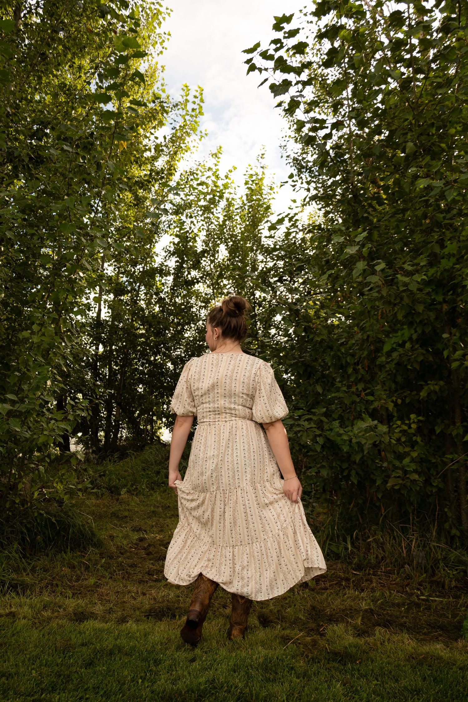 Woman in floral midi dress walking toward lush green trees on grass path in natural outdoor setting.