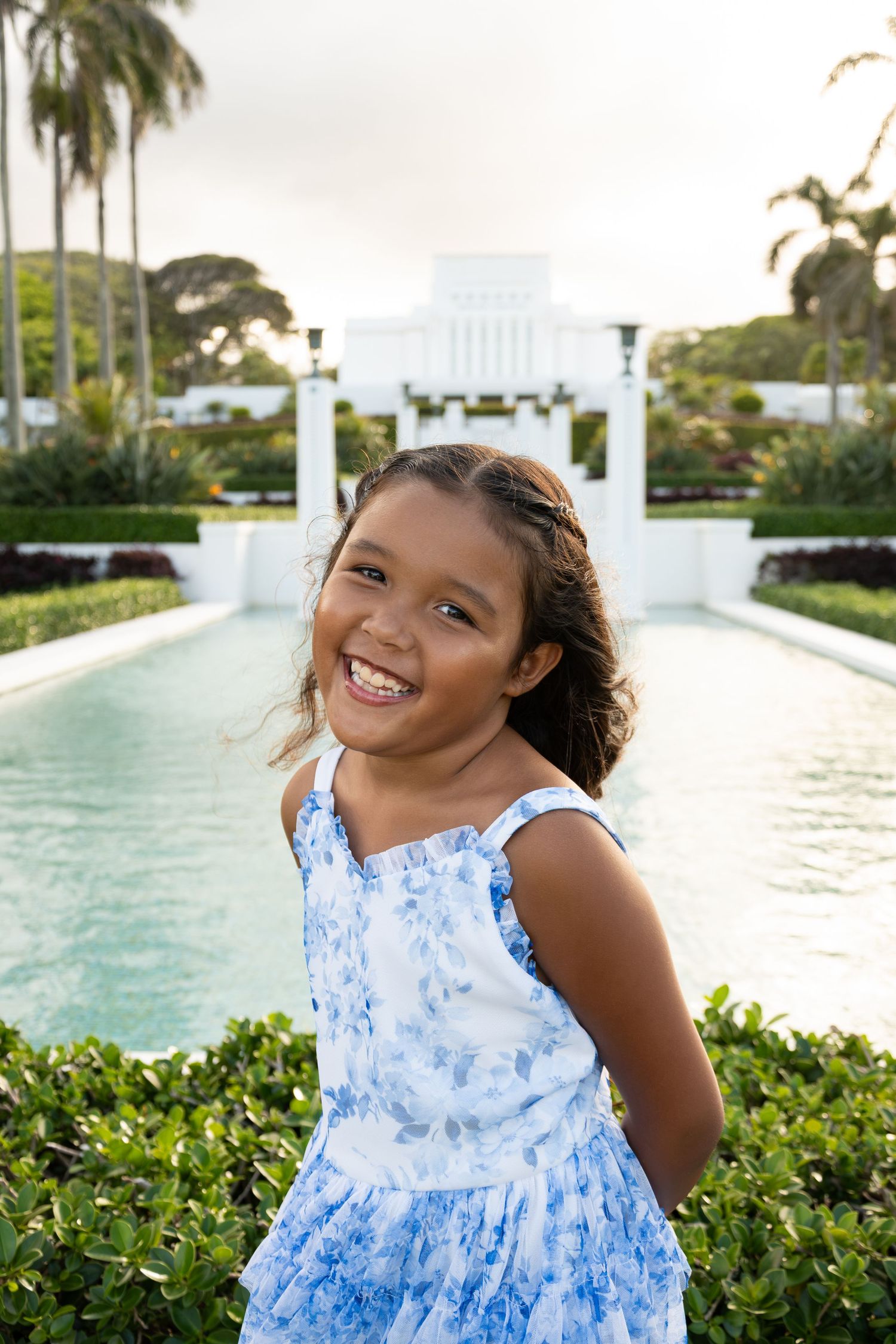 A smiling young girl in a blue floral dress stands before a reflective pool and white building surrounded by palm trees.