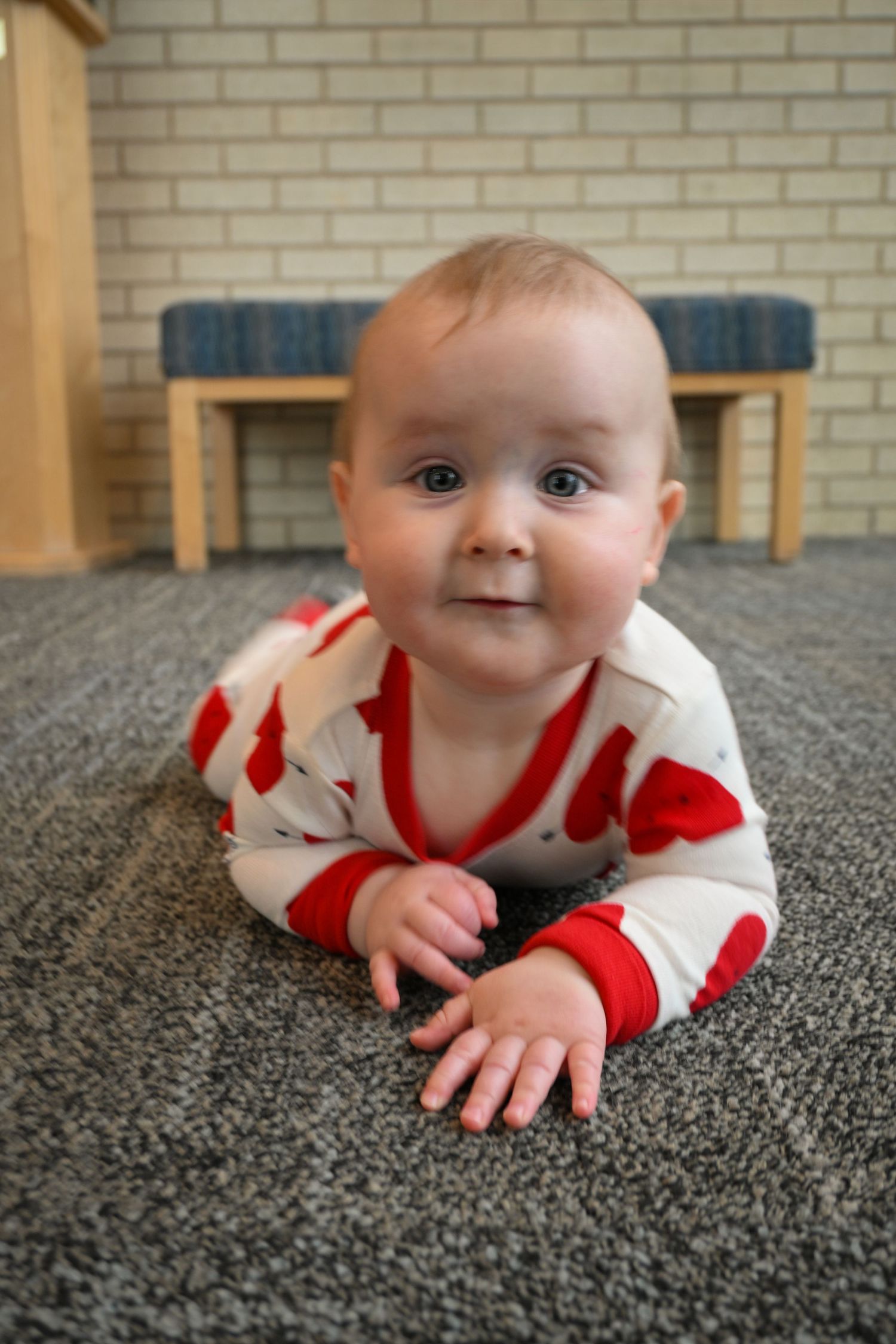 Baby in red and white heart-patterned outfit lying on gray carpet looking at camera.