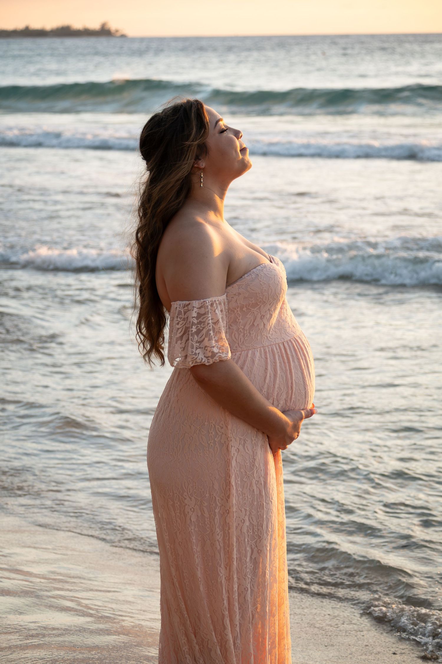 Pregnant woman in pink lace off-shoulder gown cradles her belly at sunset on a beach with ocean waves.