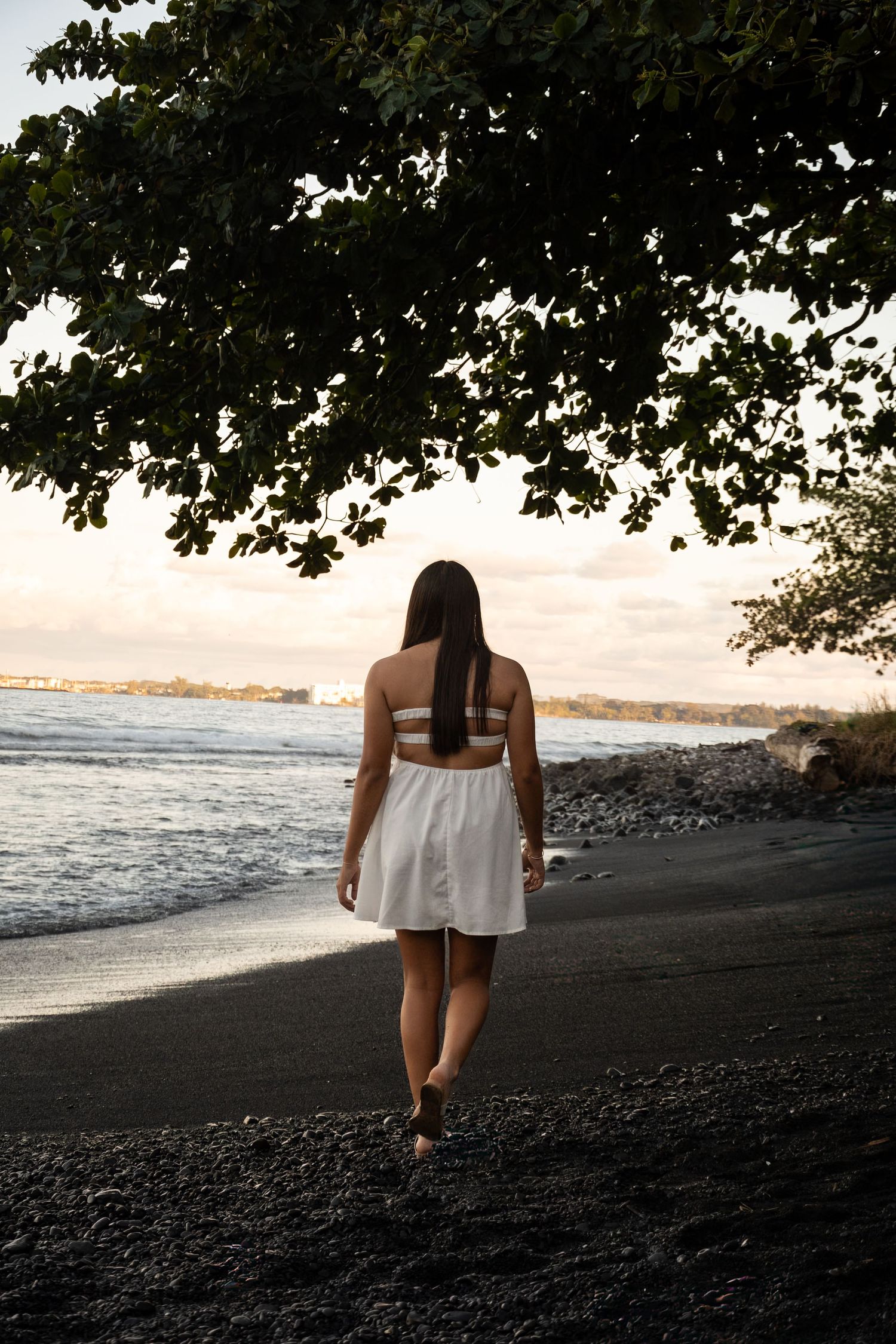Person in white dress walking on black sand beach at sunset with ocean and trees in background.