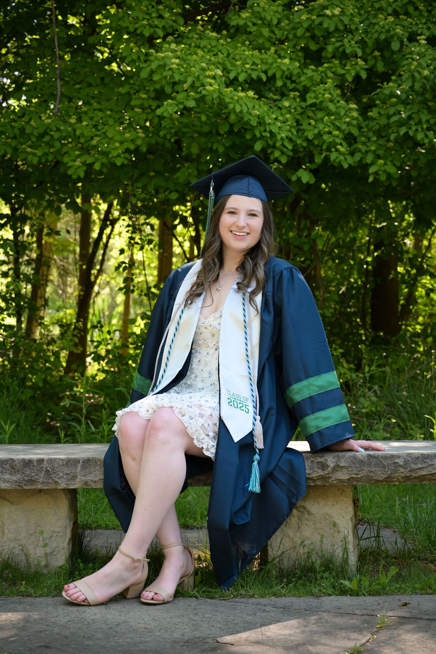 Graduate in cap and gown sits on stone wall in wooded outdoor setting during photo session.