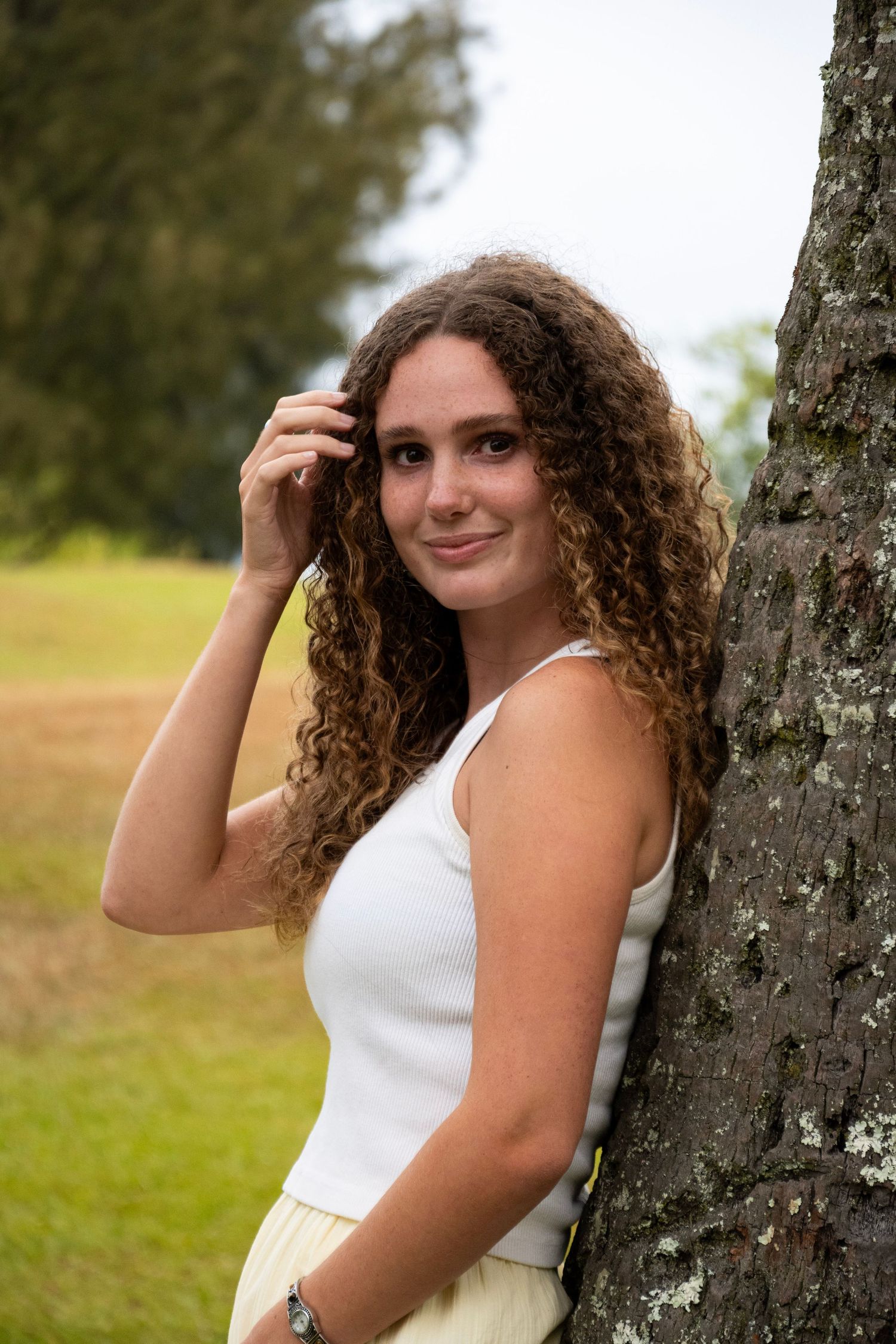 A person with curly brown hair poses against a tree trunk in a white sleeveless top during an outdoor photoshoot.