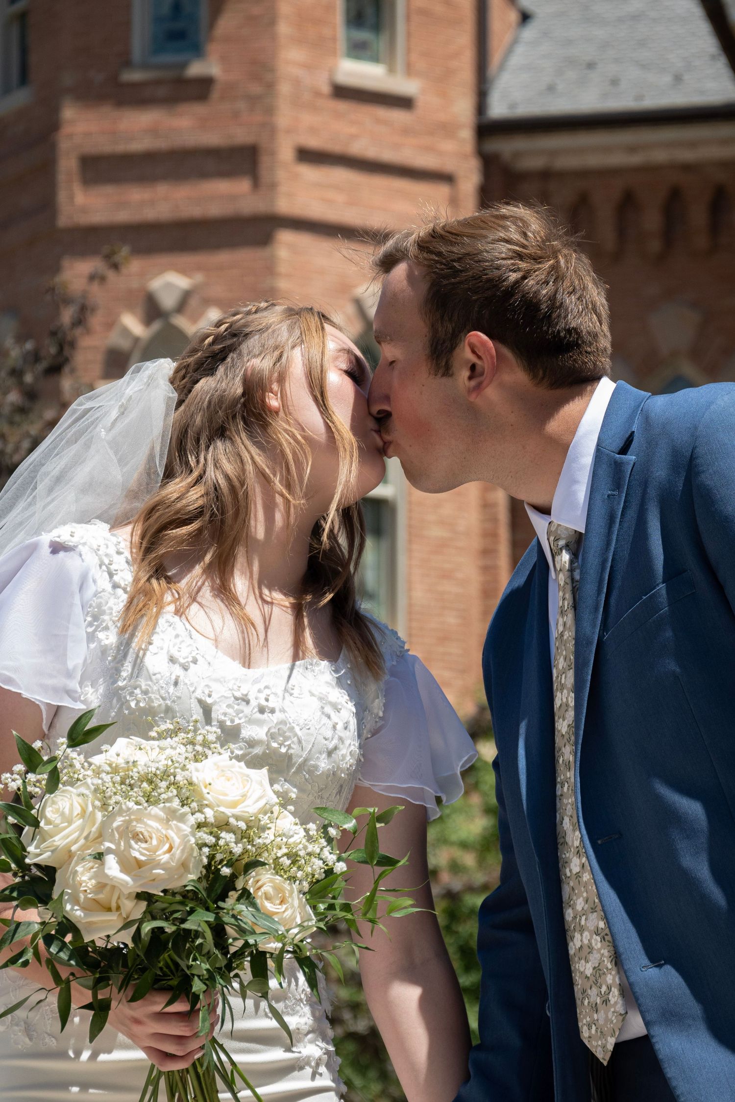 A romantic wedding couple shares a tender kiss outside a brick building while holding a white rose bouquet.