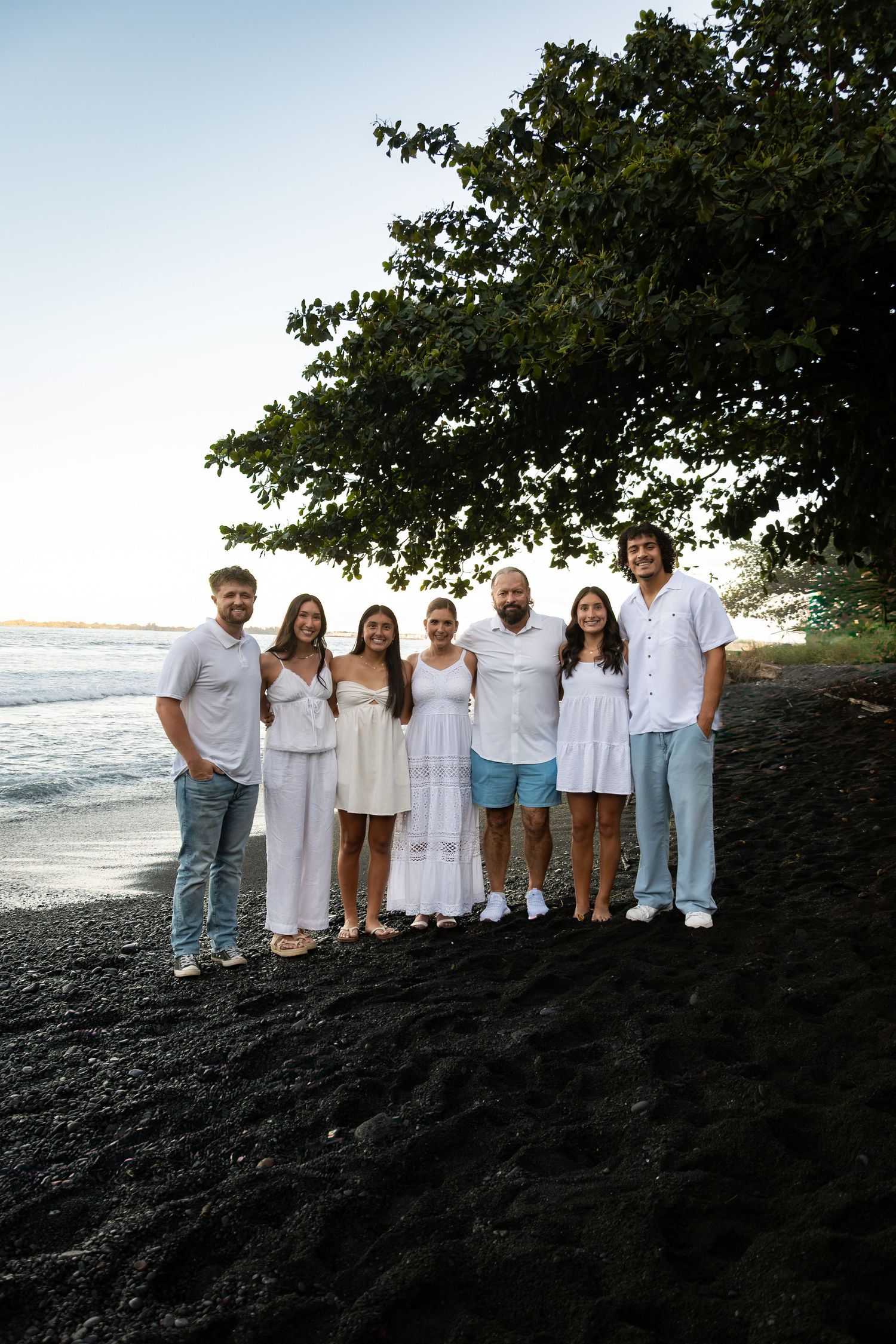 A group in white clothing stands together on a black sand beach with ocean waves and a tree overhead.