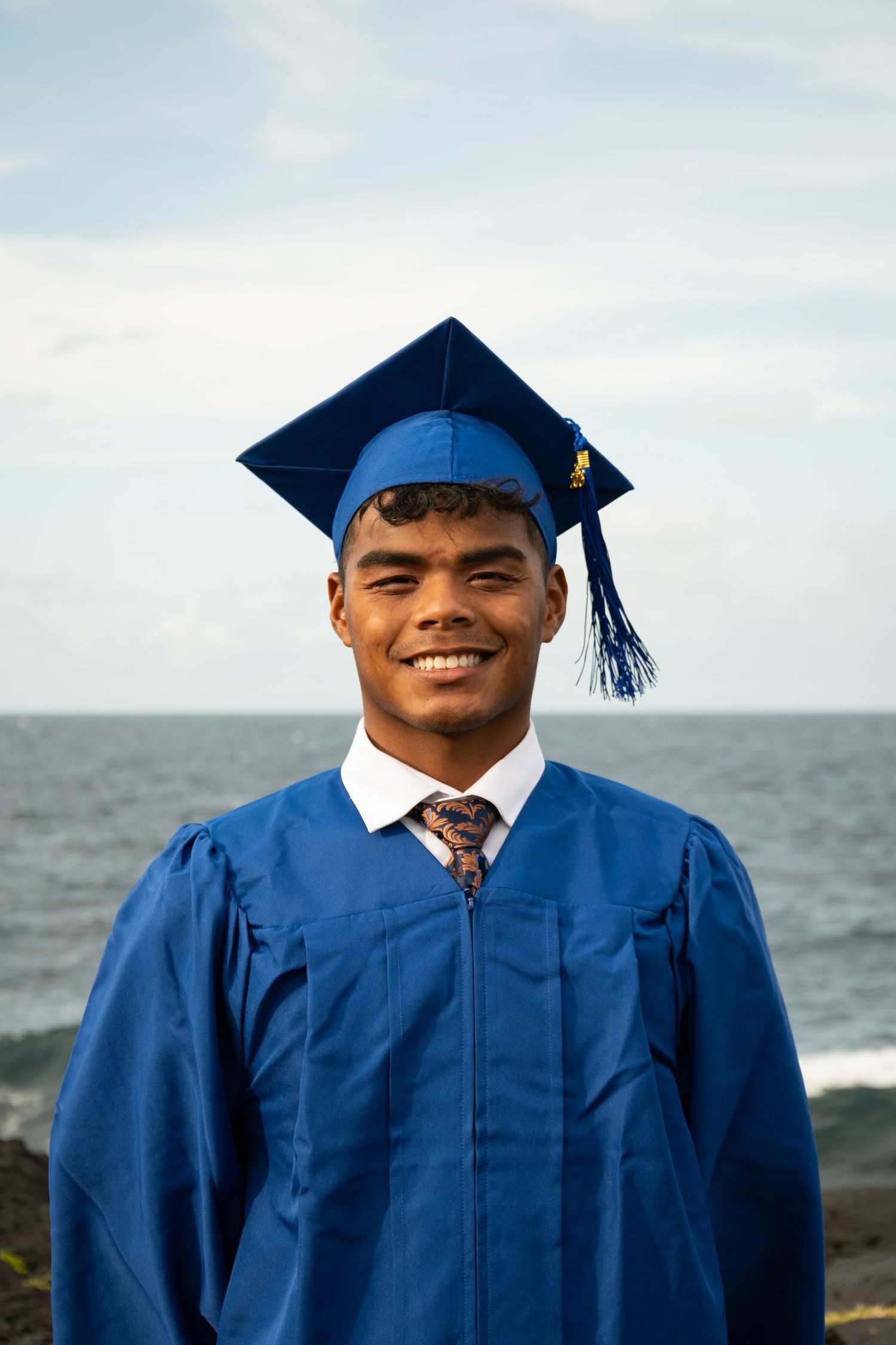 Smiling young man in blue graduation cap and gown posing outdoors by the ocean on a cloudy day.