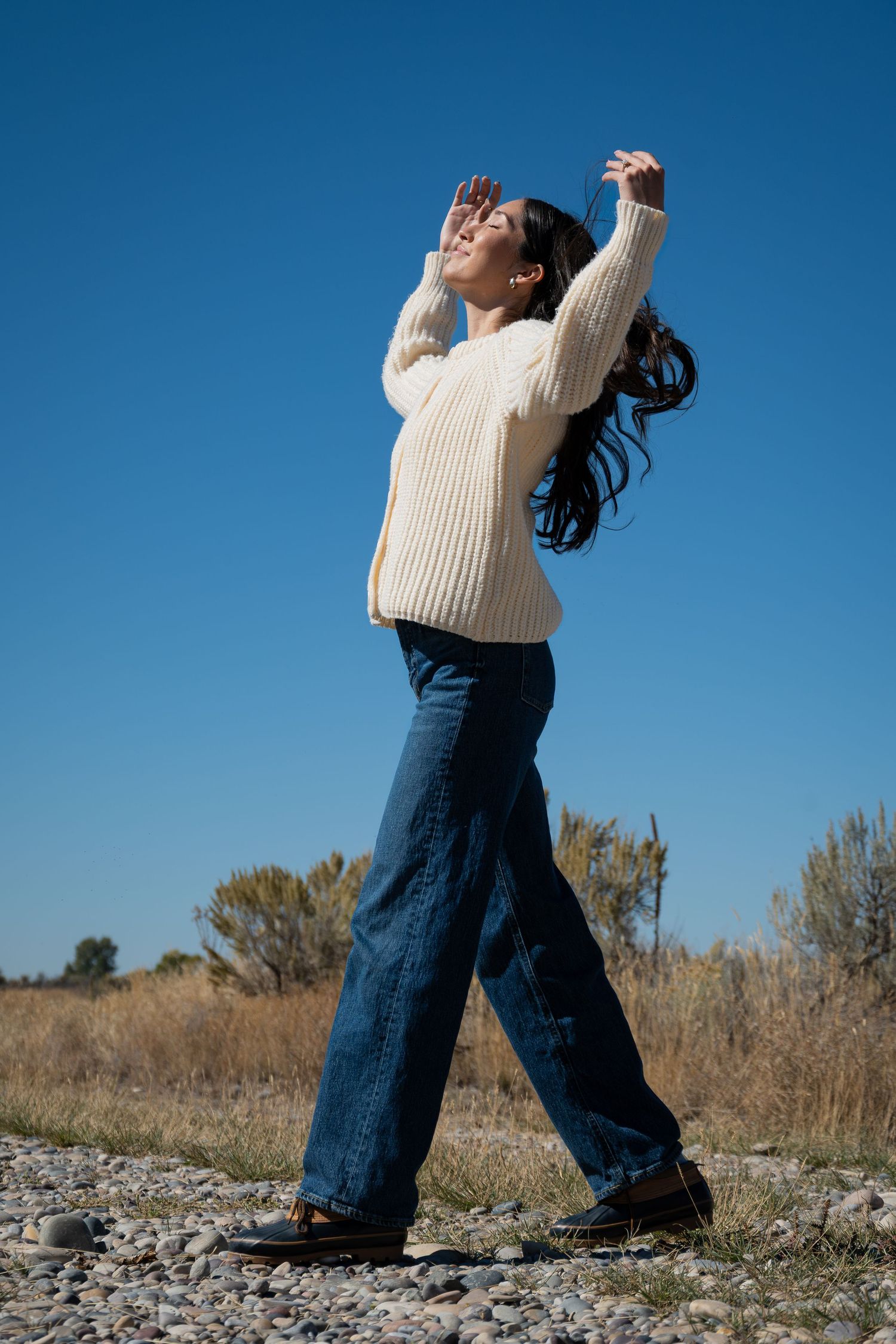 Someone in a cream sweater and wide-leg jeans dances on rocky terrain against a bright blue sky background.