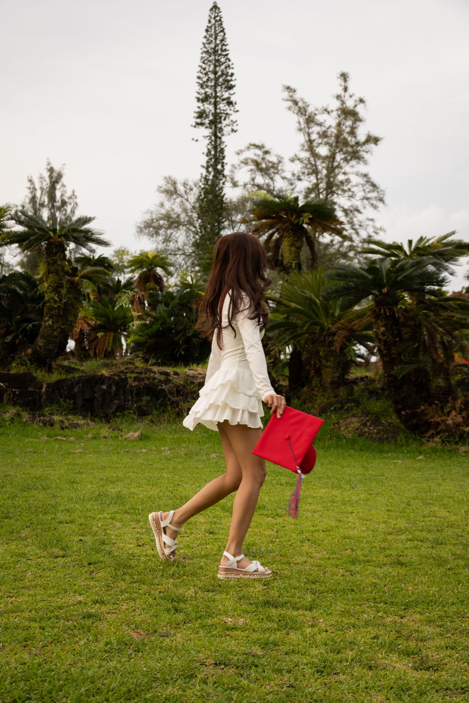 Young girl in white dress holding red graduation cap walks across lush green lawn with tropical trees.