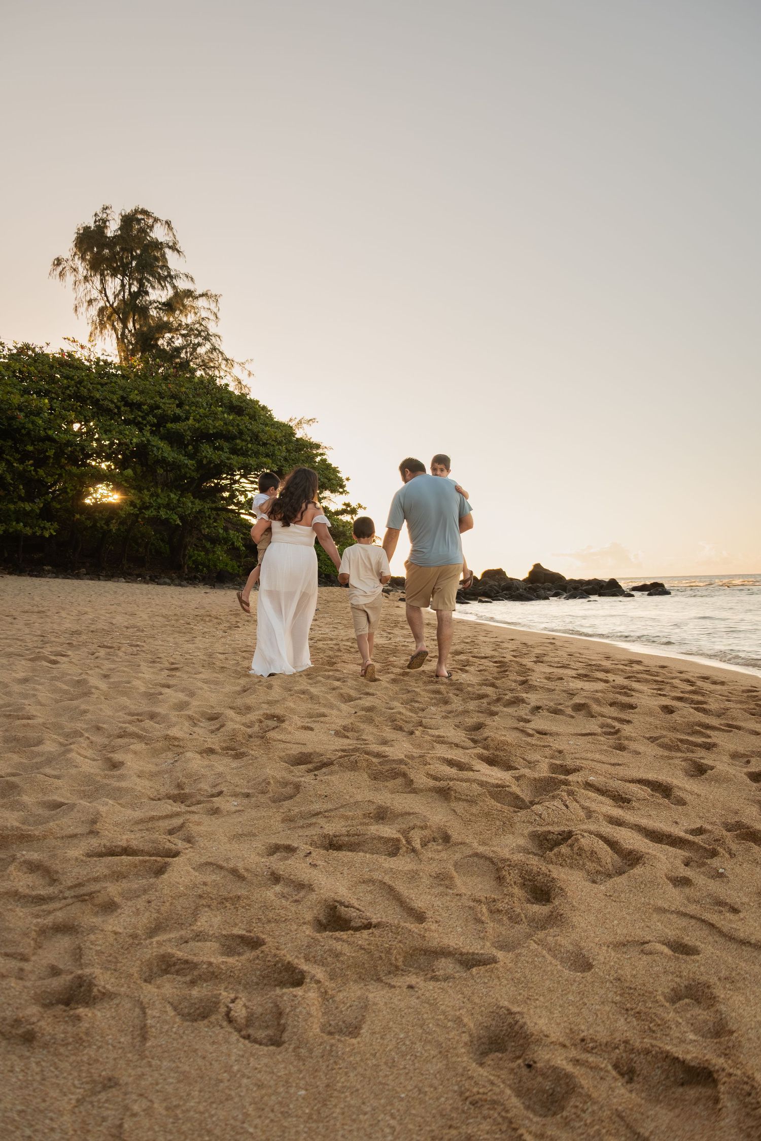 Family walking on sandy beach at sunset, leaving footprints in sand near tropical trees and ocean waves.