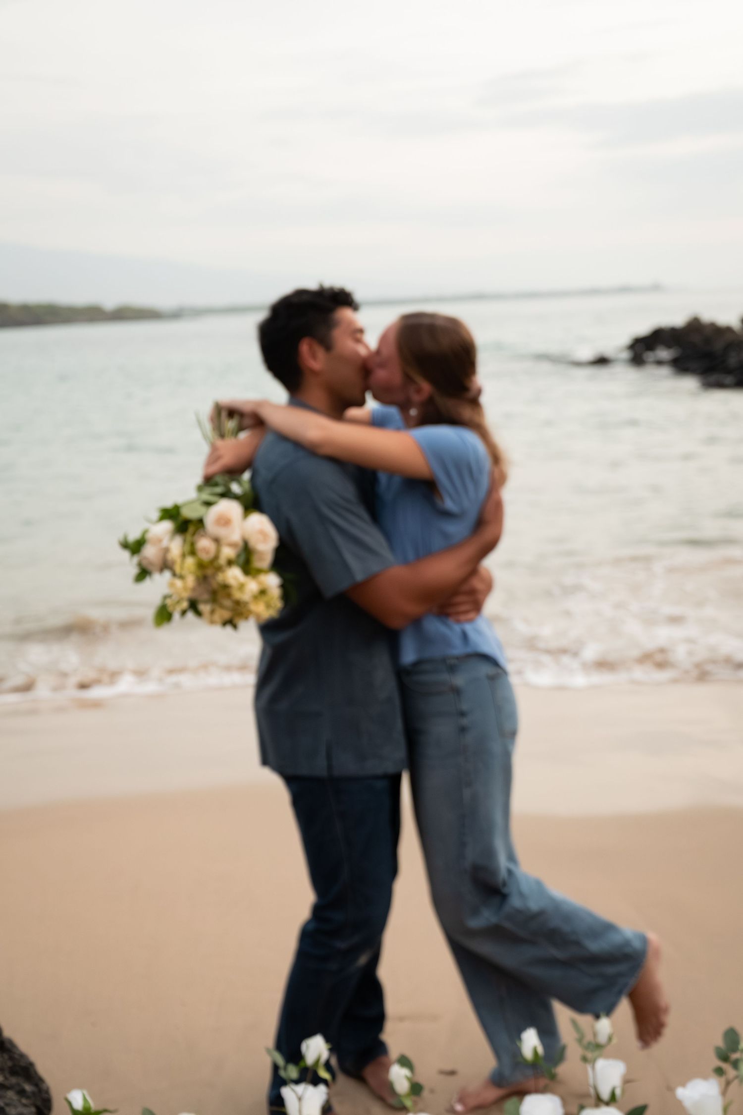 Couple kissing on a beach while holding a white flower bouquet with roses scattered on the sand near the ocean.
