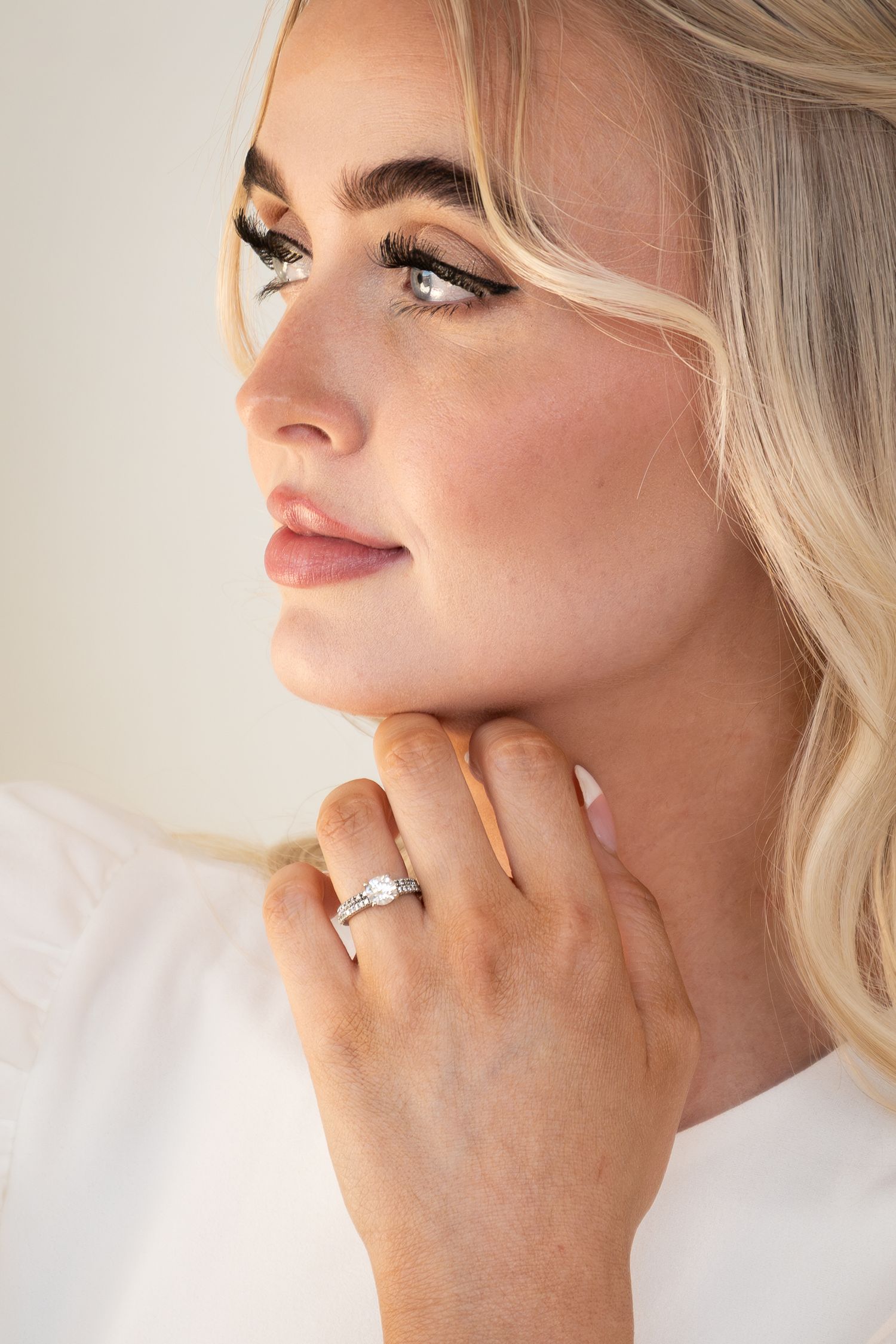 Blonde woman wearing a diamond solitaire engagement ring, touching her chin and smiling softly in a white top.