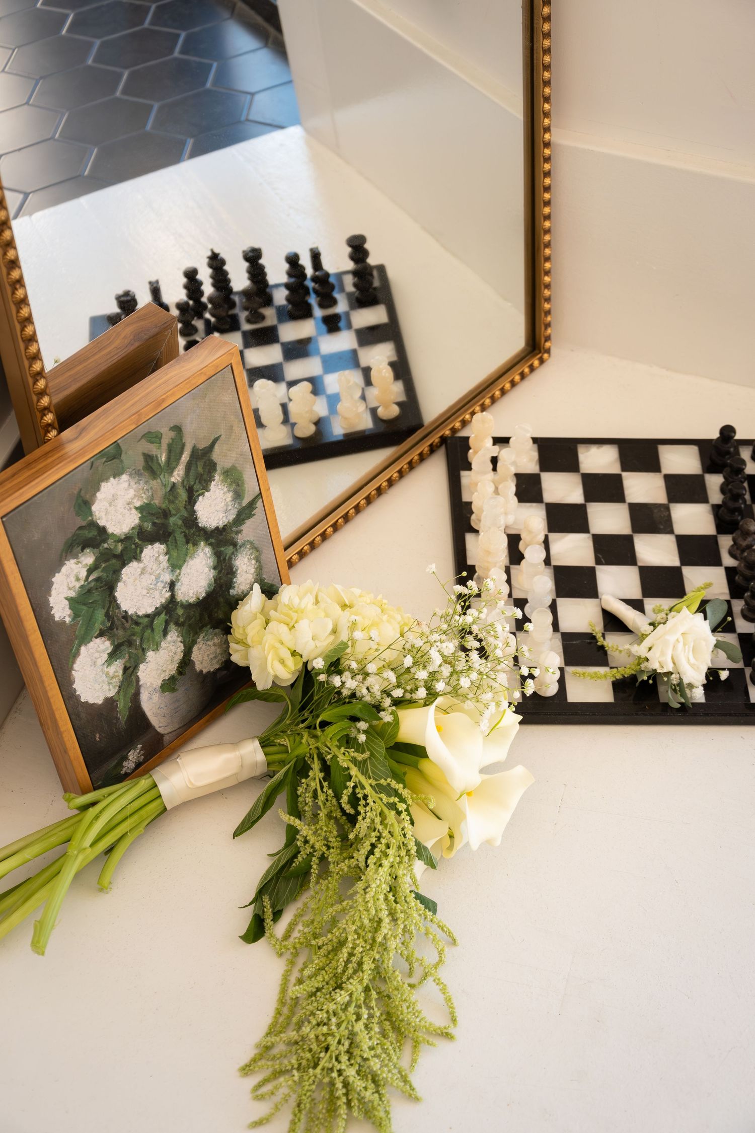 Elegant wedding flat lay with white rose bouquet, chess board, floral painting, and gold mirrors on white surface.