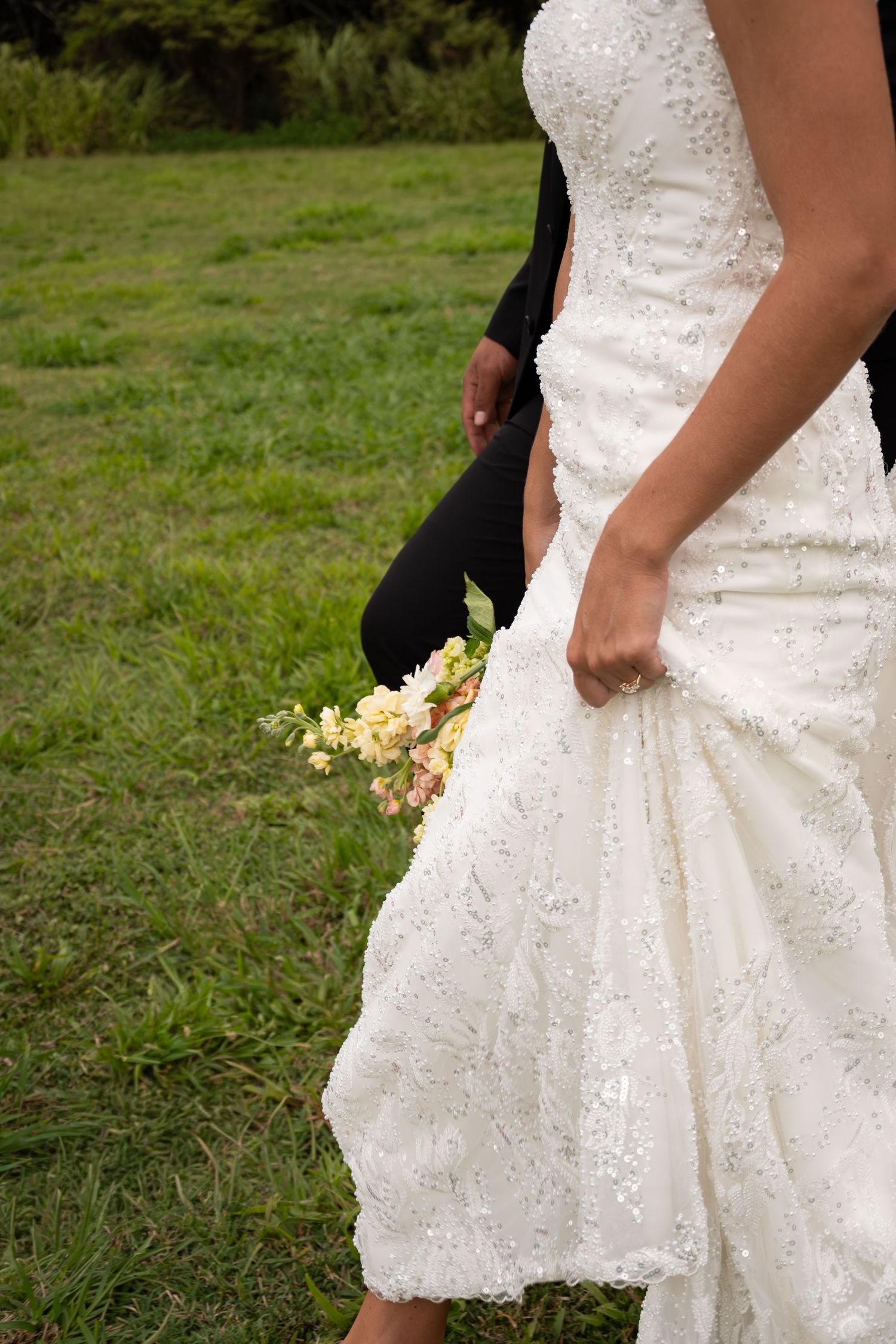 Close-up of bride in beaded white gown holding bouquet of pastel flowers while walking with groom.
