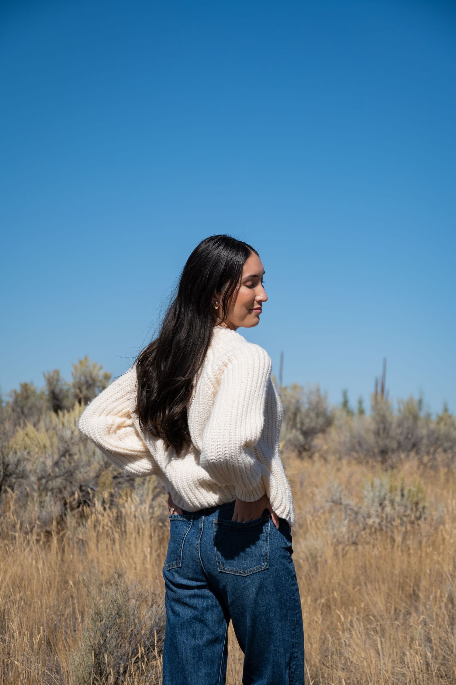 Person wearing white knit sweater and jeans stands in field of dry grass against bright blue sky.