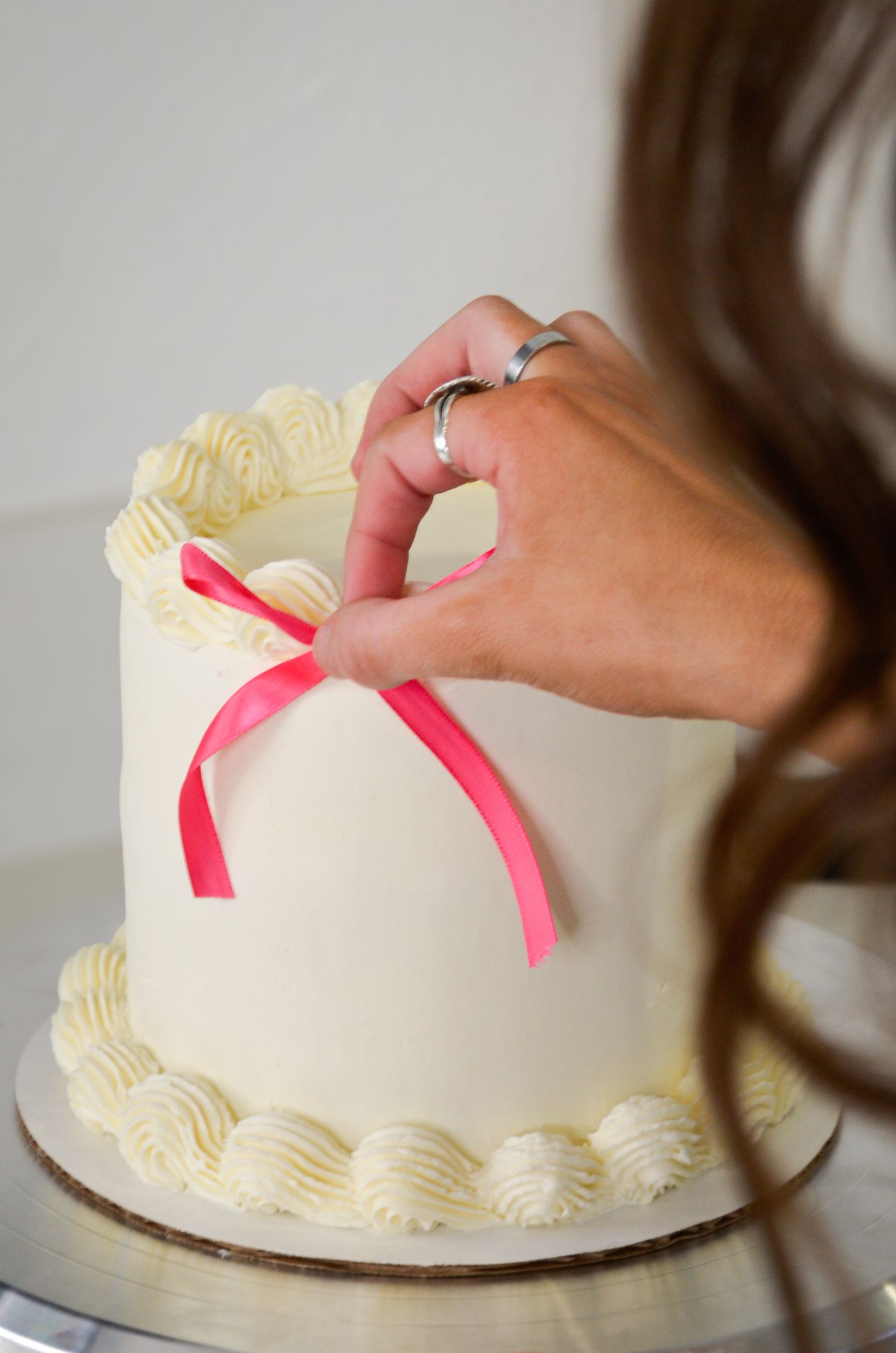 Hand with wedding ring tying pink ribbon on white buttercream cake.