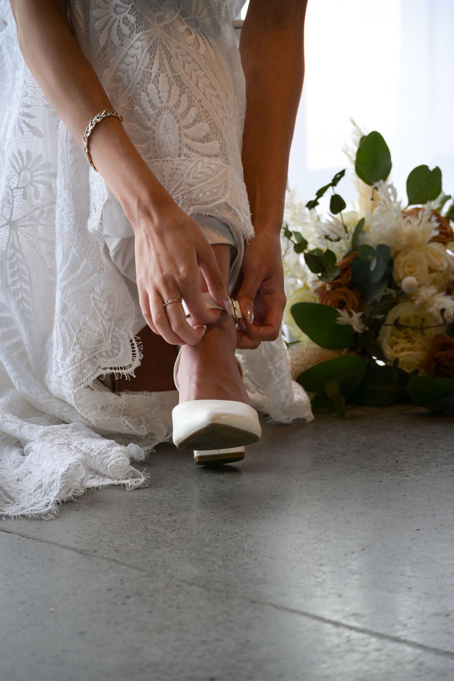 Close-up of bride putting on white wedding shoes with lace dress detail.