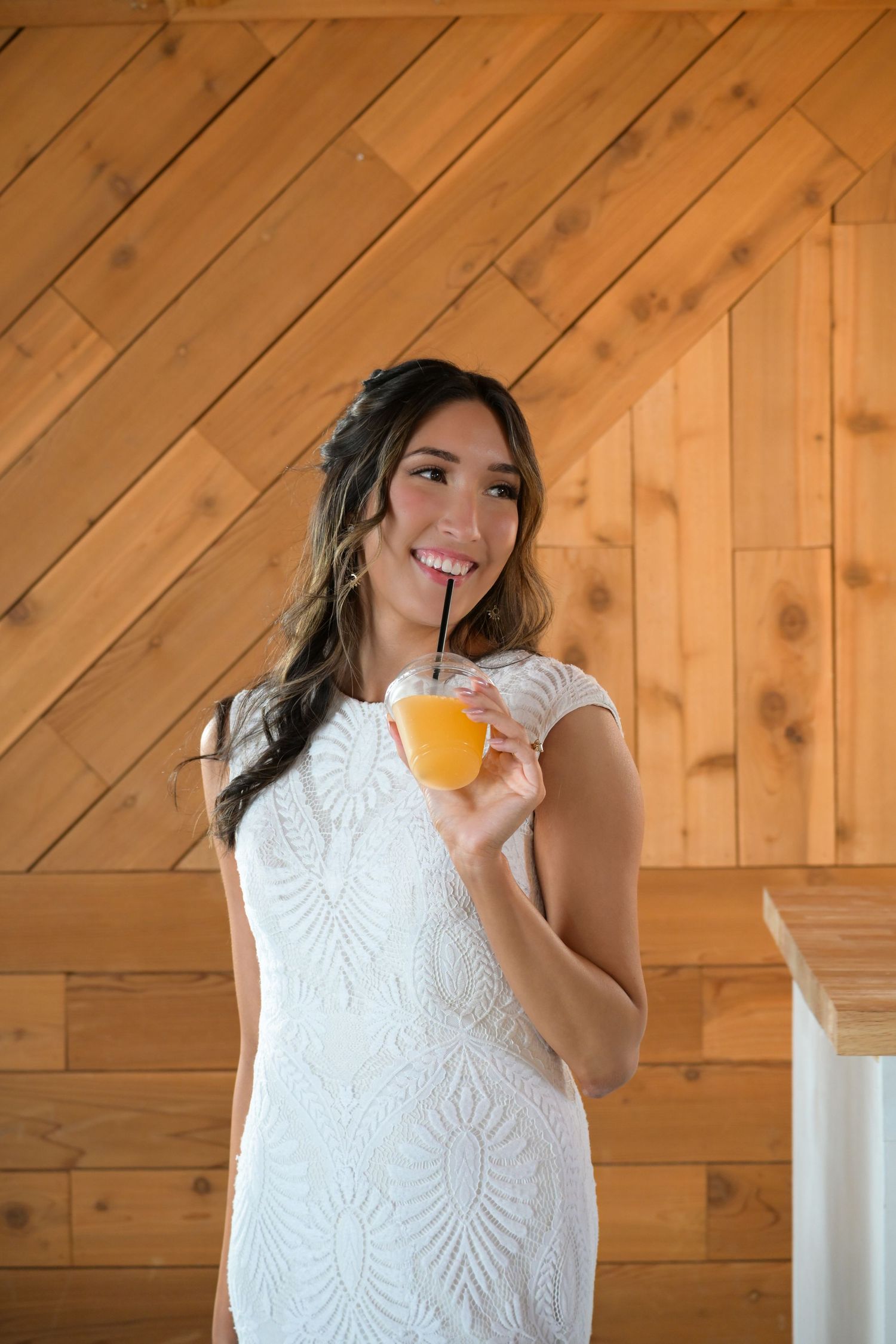 A bride in a white lace dress sips from an orange drink through a straw.