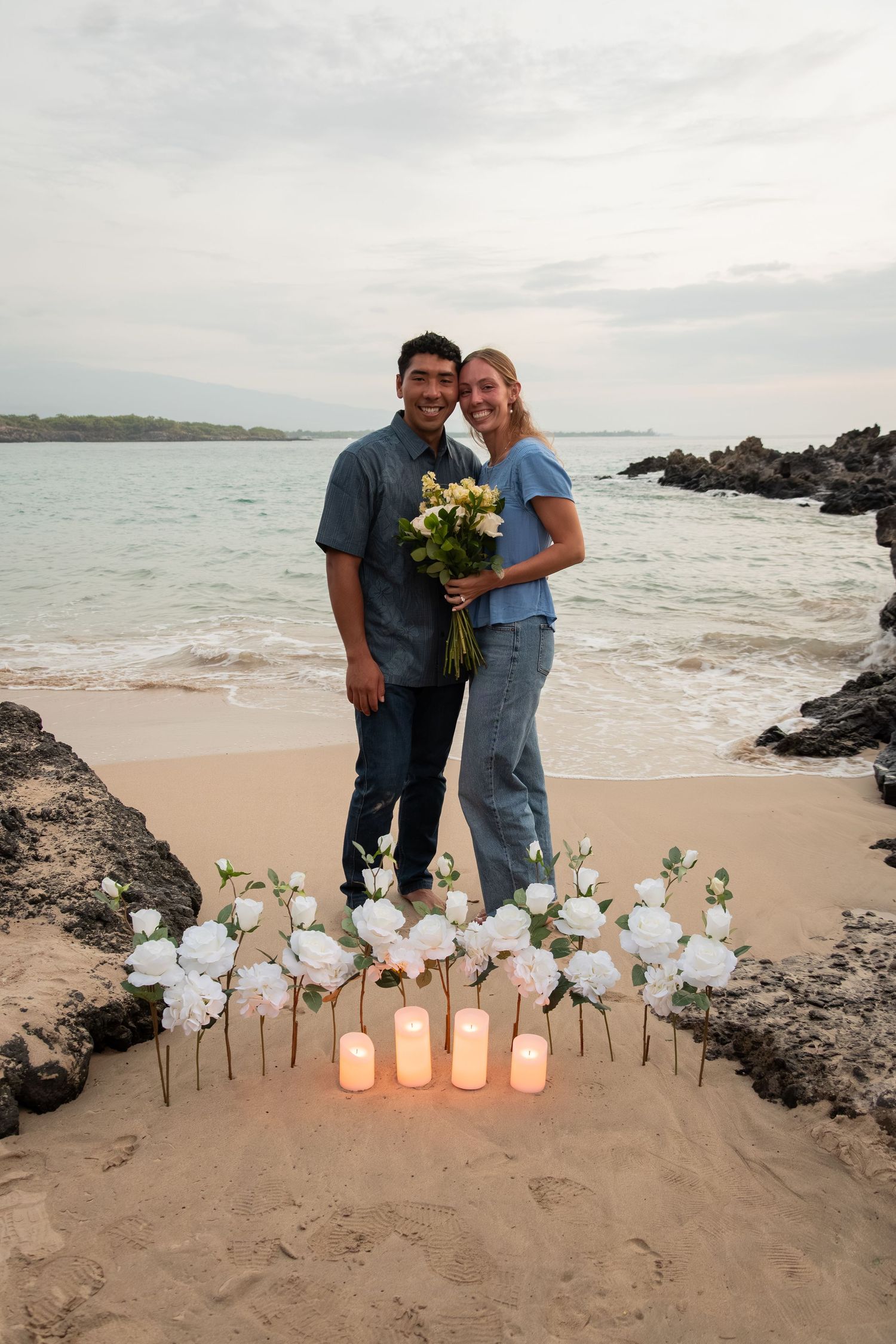 Couple smiling on a beach at sunset with white roses and candles arranged in the sand during a romantic proposal.