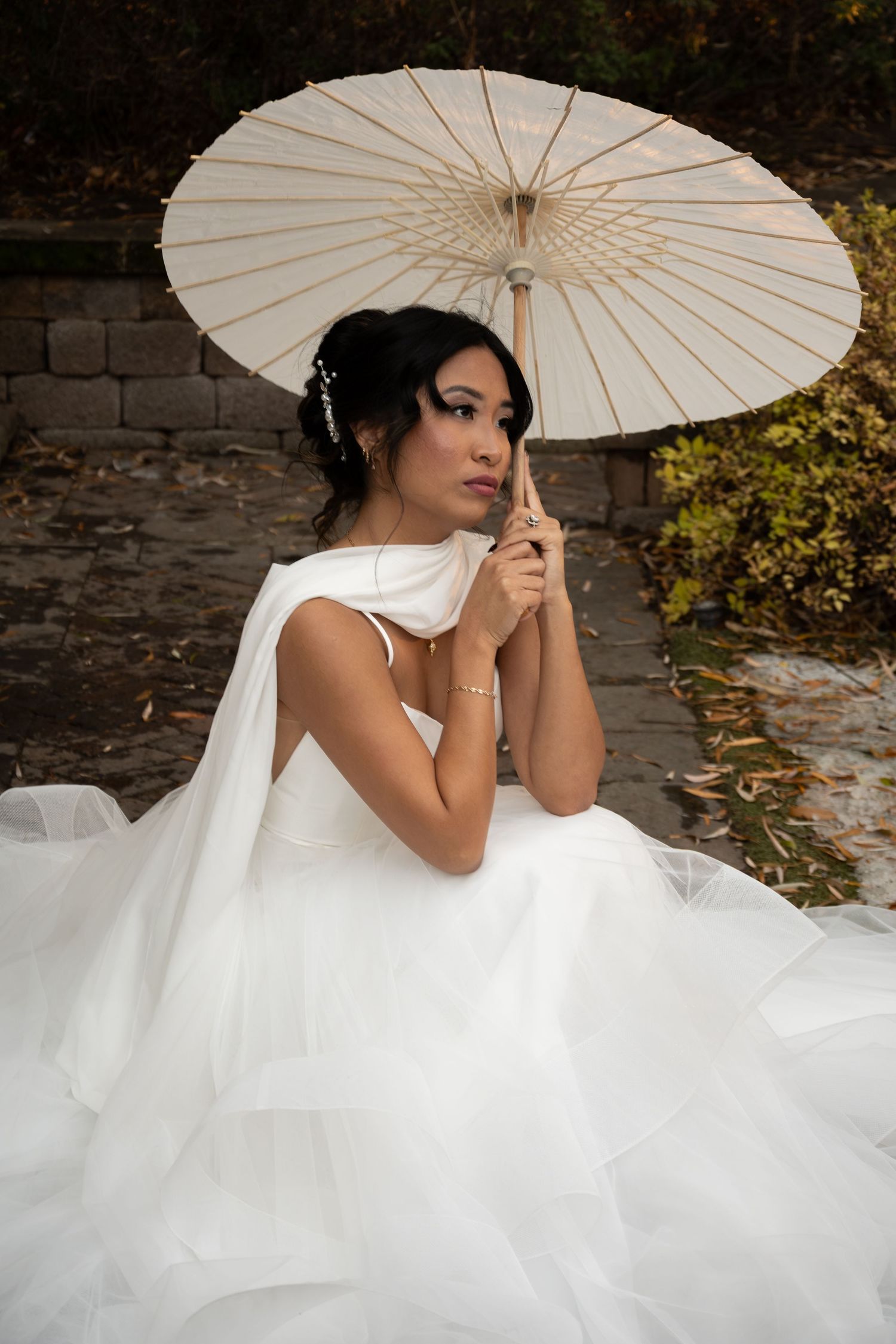 Elegant bride in white tiered gown holding a traditional paper parasol umbrella outdoors in a garden setting.