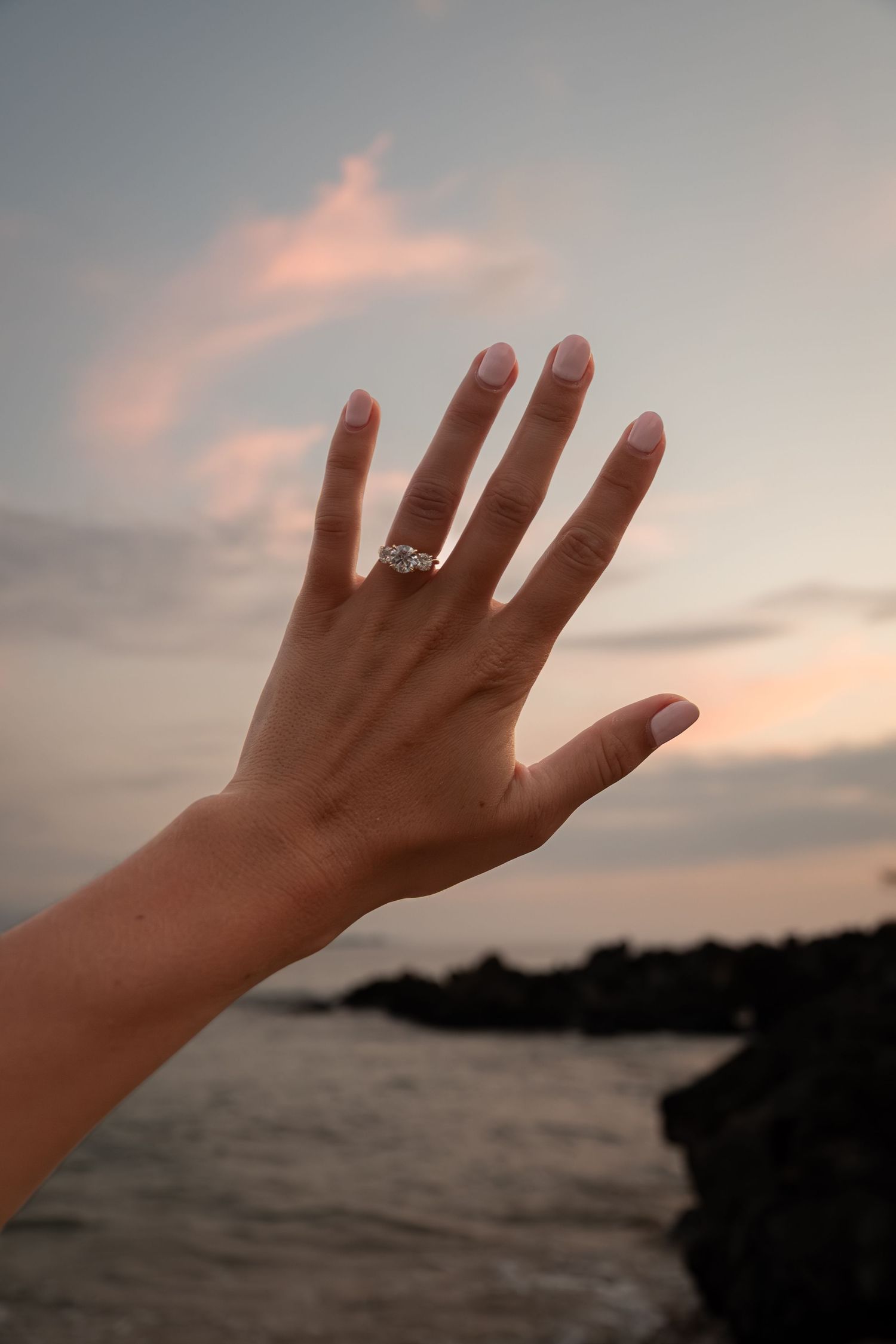 A hand with a diamond engagement ring raised against a scenic ocean sunset backdrop with rocky shores.
