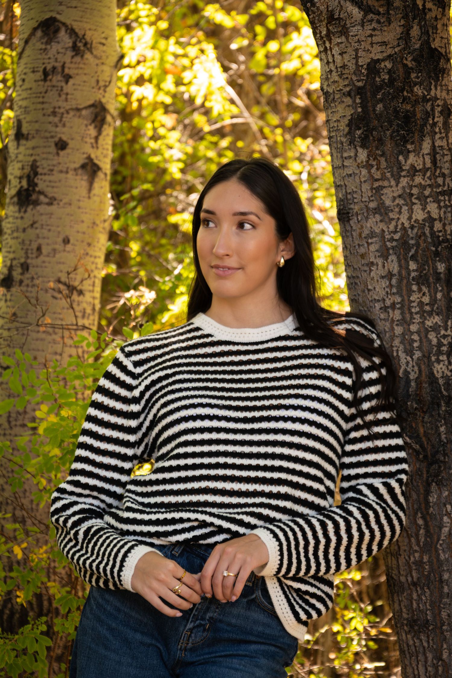 Person wearing black and white striped sweater poses casually in a sunlit forest setting with birch trees.