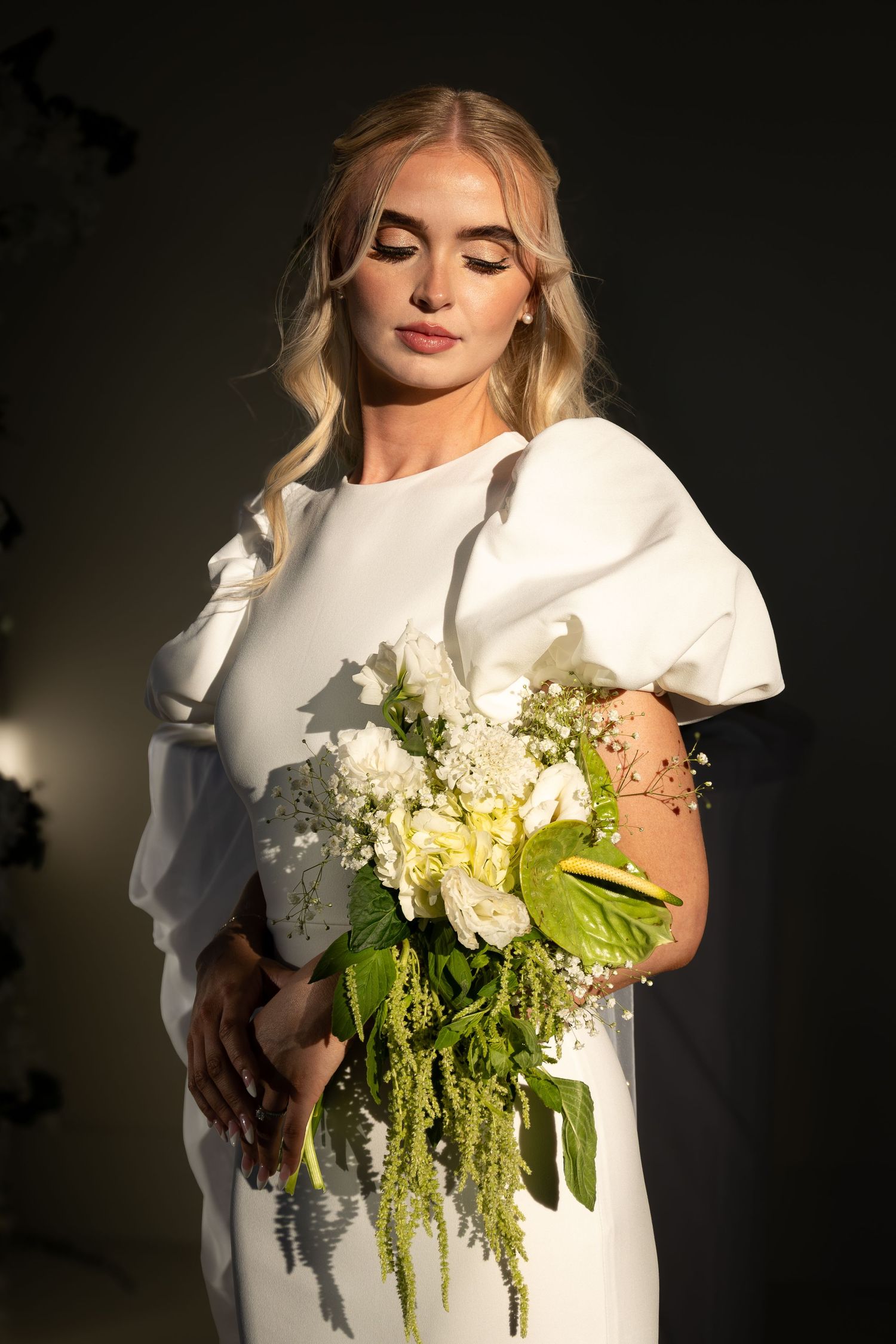 Elegant bride in white dress with puff sleeves holding a bouquet of white and green flowers, looking down gracefully.
