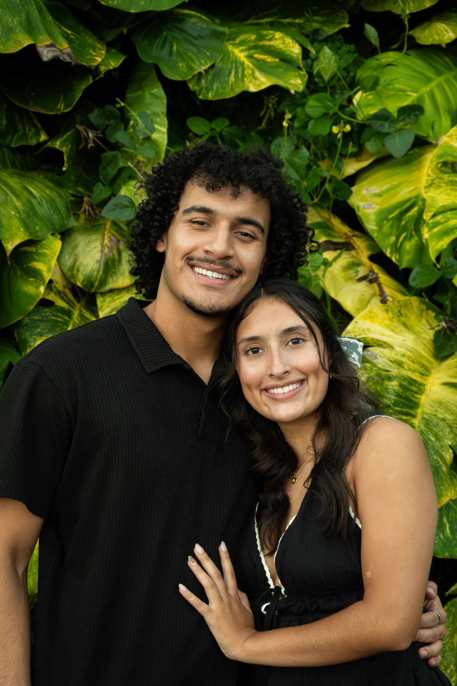 A couple poses together against lush green foliage in a romantic outdoor photoshoot.