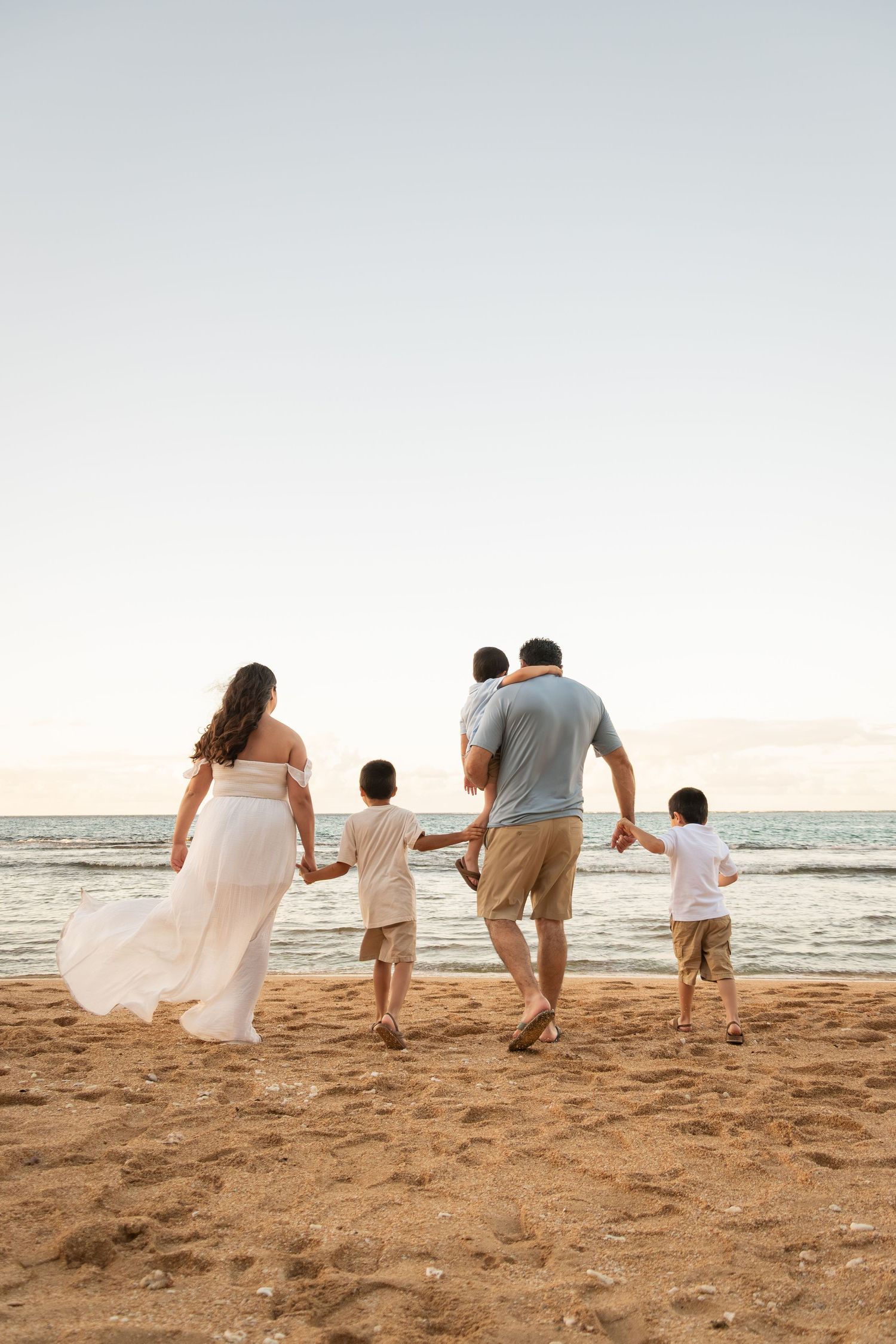 Family of four walking hand-in-hand on a sandy beach at sunset, with two boys and a woman in a flowing white dress.