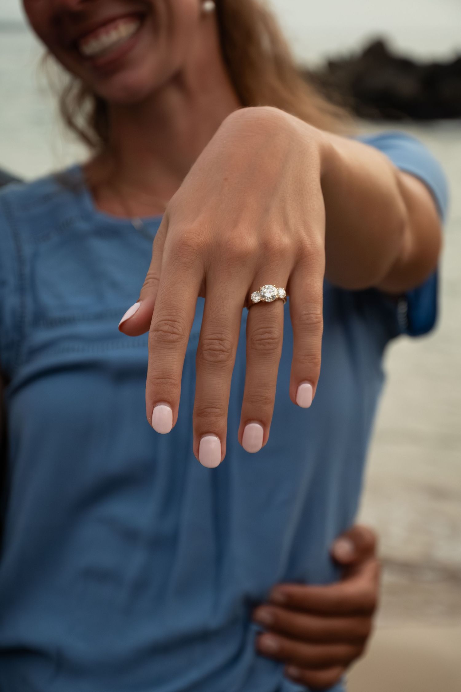 Woman in blue shirt showing off diamond engagement ring with pink nails at beach.
