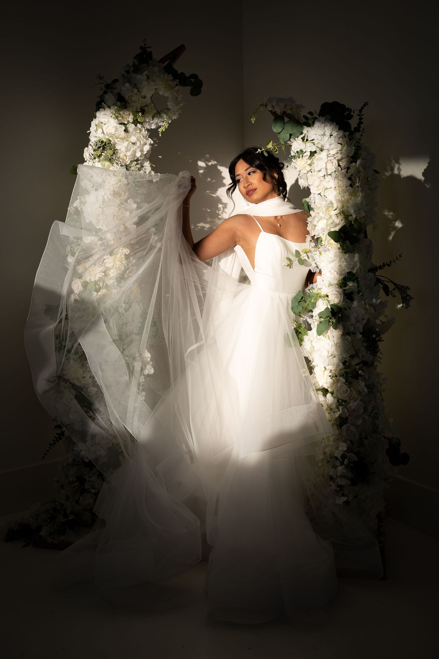 Elegant bride in a white tulle gown posing between two floral arches with white blooms.