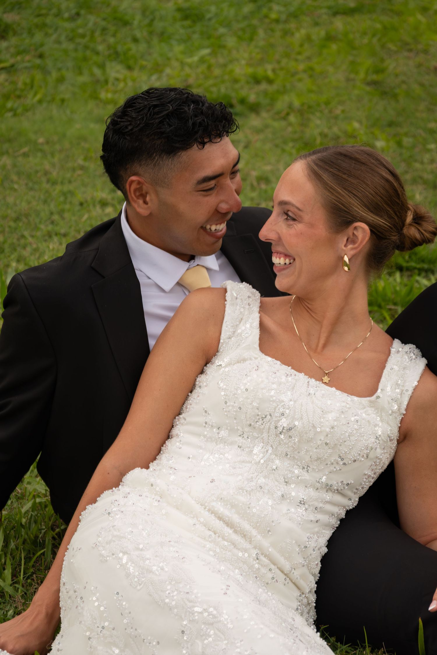 Smiling bride and groom in black suit sit together on green grass sharing a joyful moment.