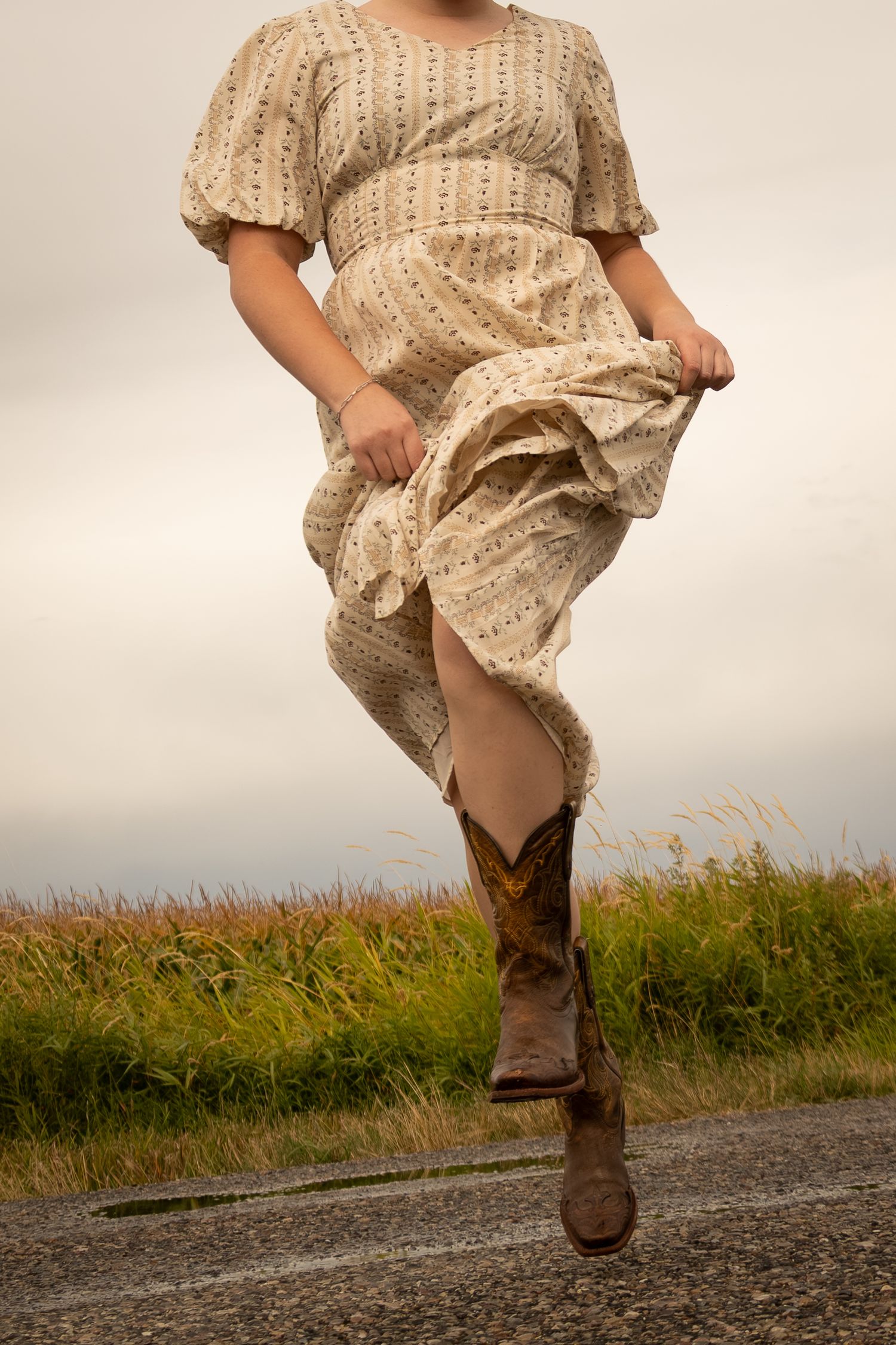Woman in floral midi dress and cowboy boots walking outdoors on a cloudy day near a grassy field.