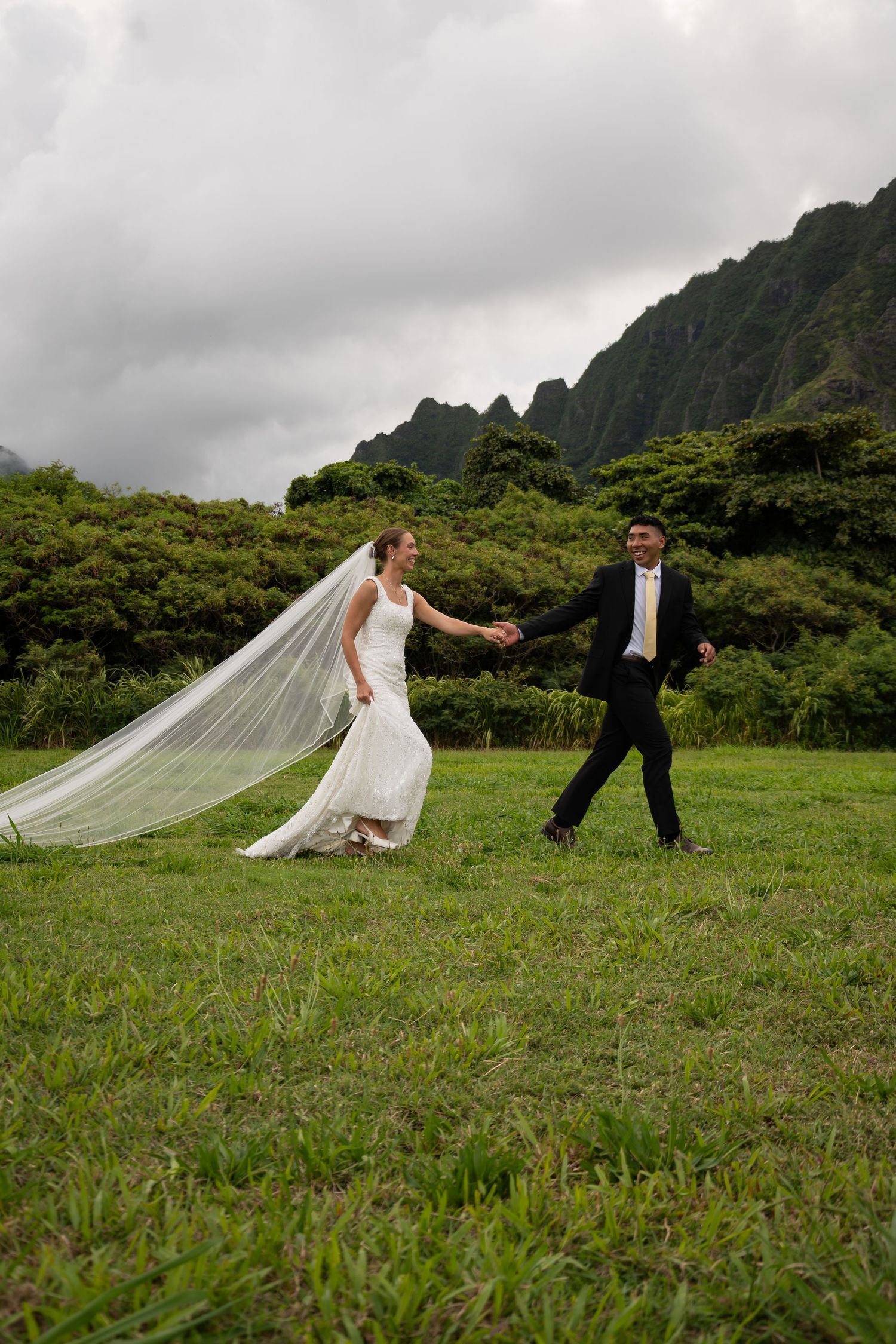Bride and groom holding hands walking on green grass with dramatic mountain backdrop on a cloudy day.