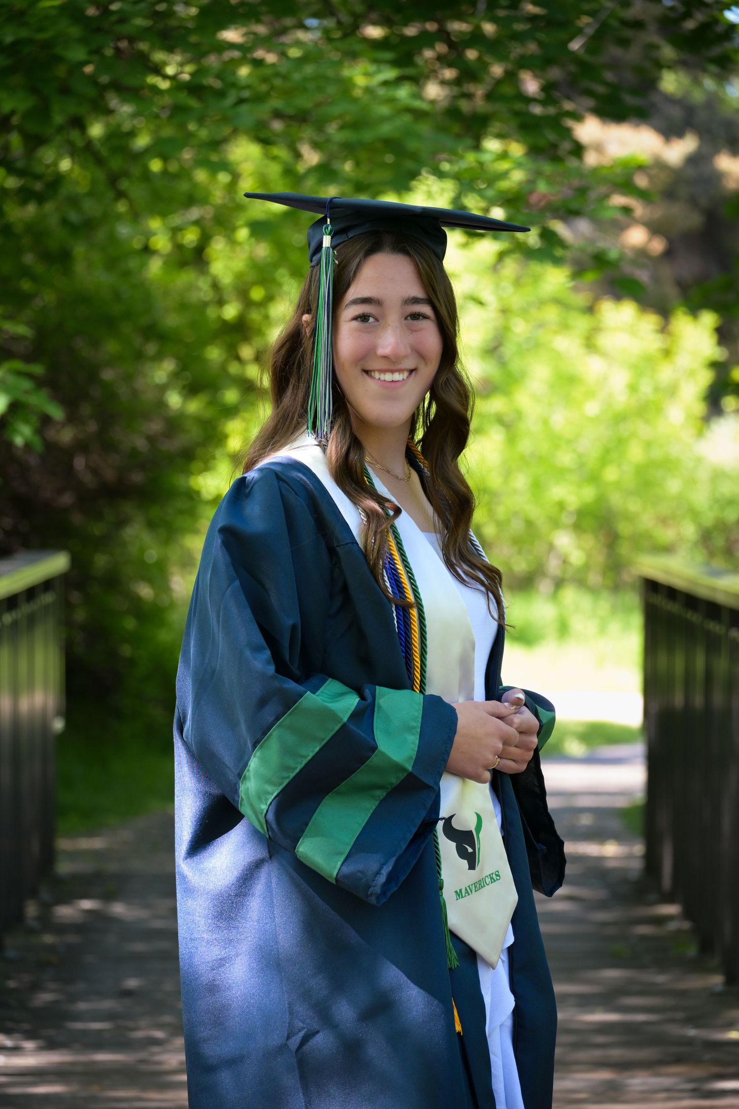 A graduate in blue cap and gown poses outdoors on a wooden walkway surrounded by lush green foliage.