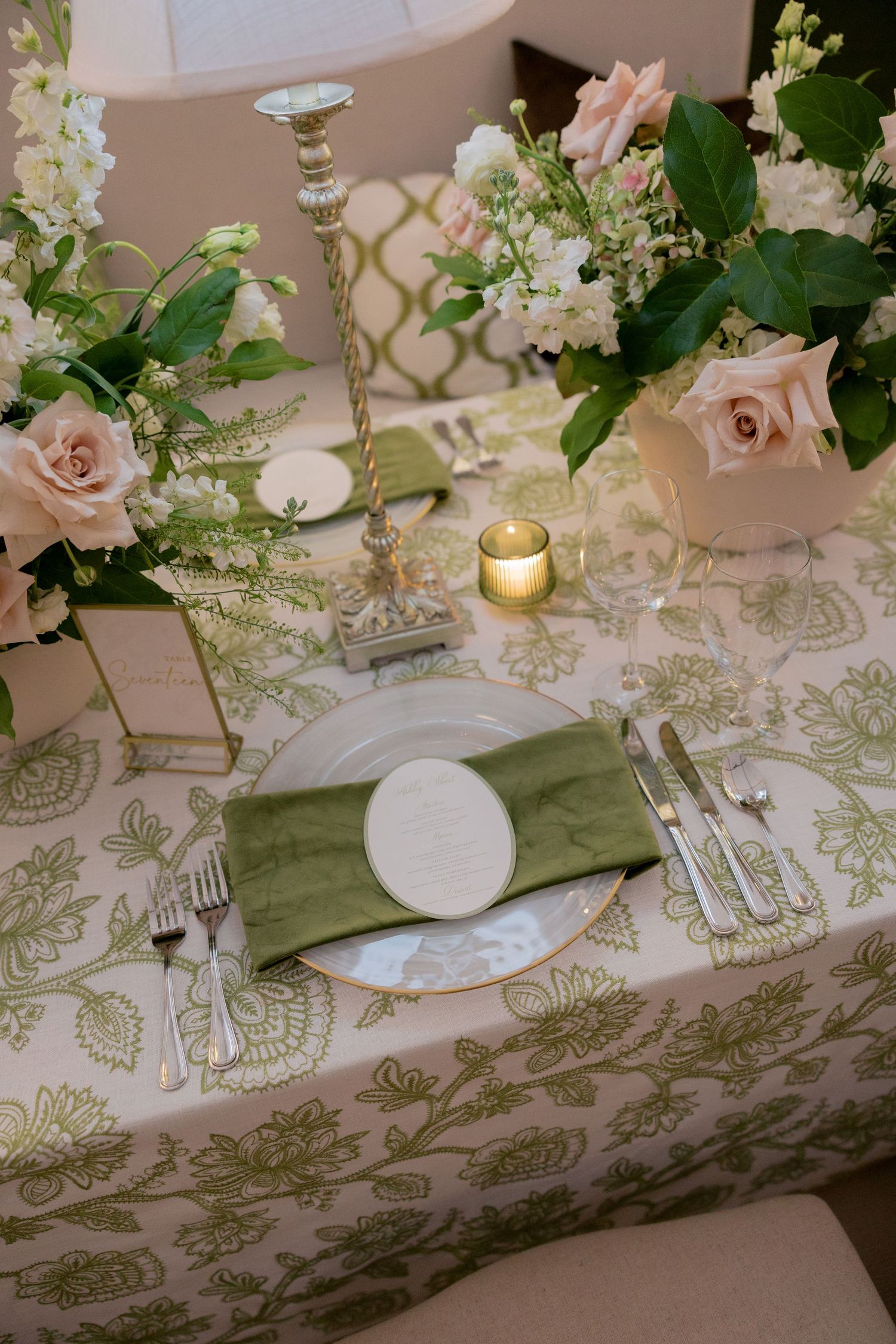 Elegant wedding table setting with green floral tablecloth, blush roses, silver candlestick, and sage green napkin on gold-rimmed plate.