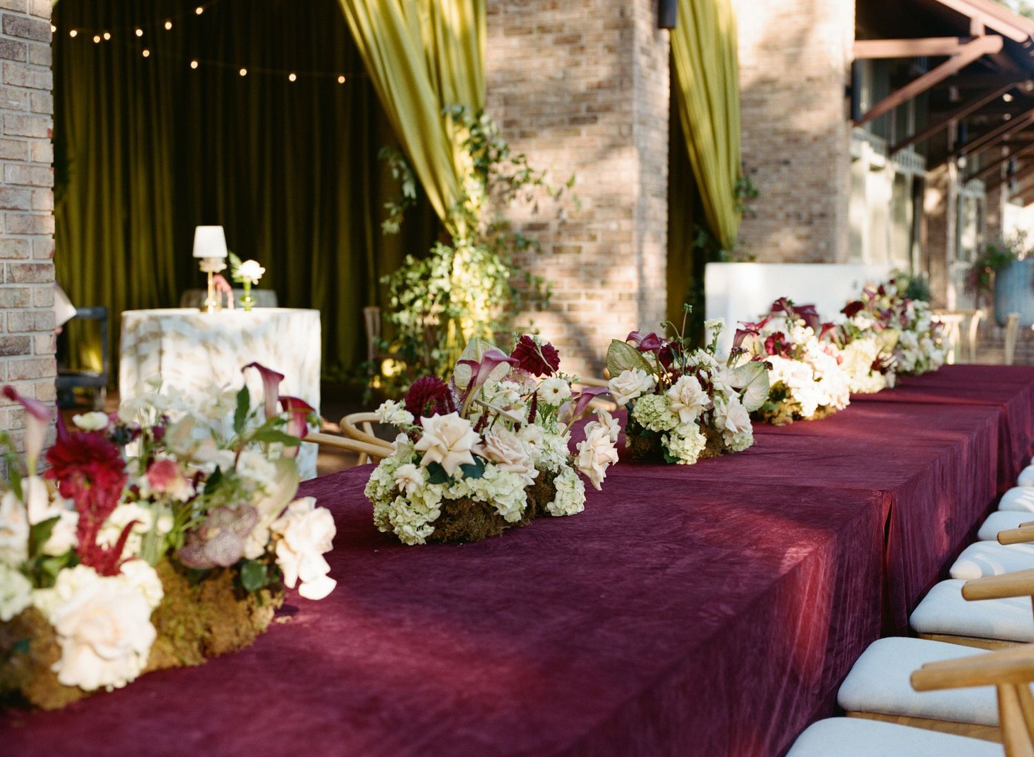 Wedding reception table draped in burgundy velvet with white and burgundy floral centerpieces, gold curtains and a rustic brick wall behind.