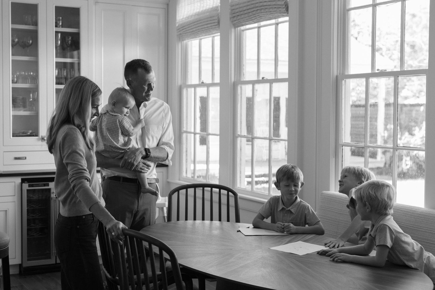 Black and white photo of a family in a bright kitchen, parents standing with a baby while three children sit at a wooden dining table.