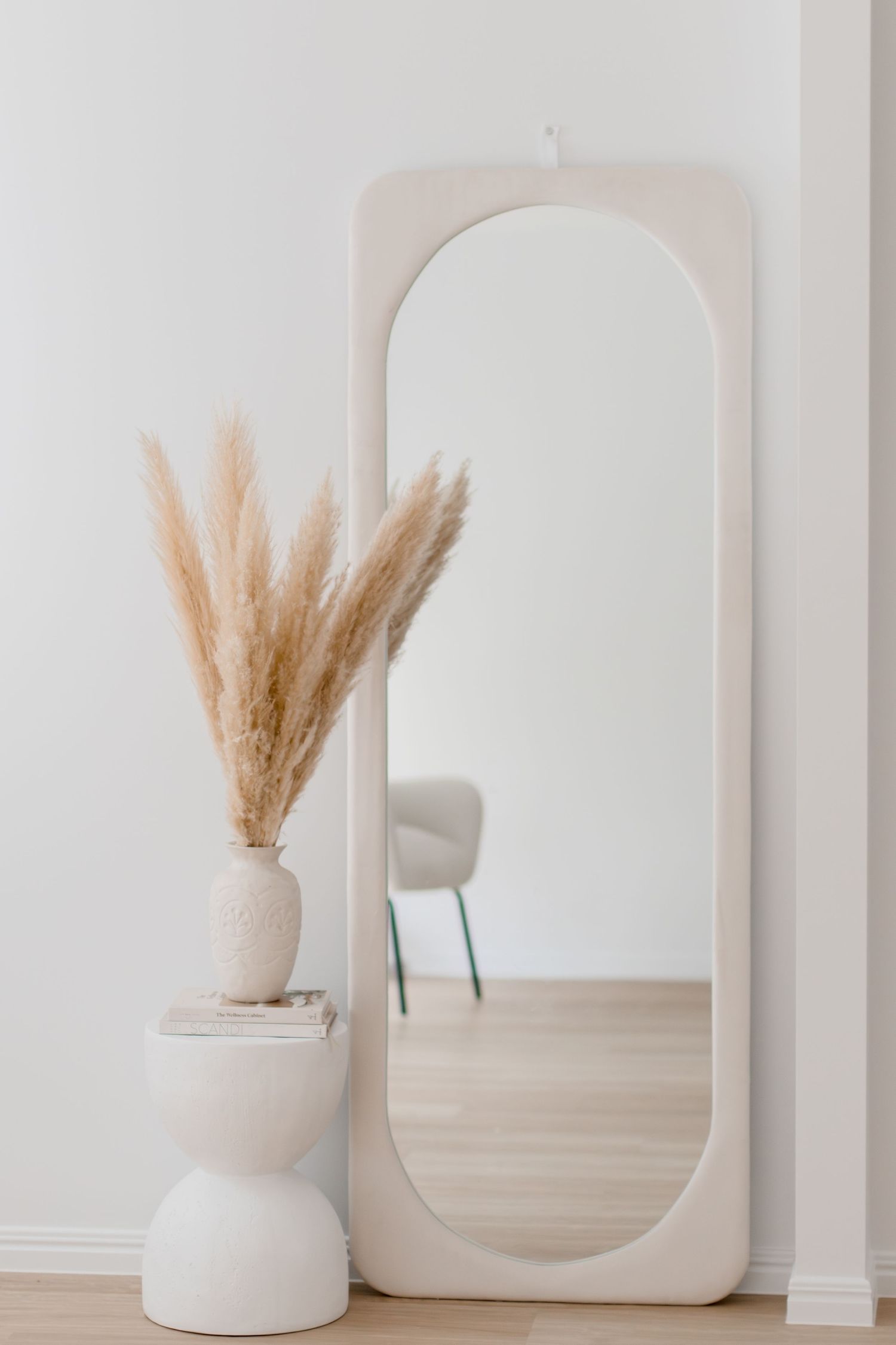 Minimalist white ceramic vase with dried pampas grass against a white wall with arched mirror creating a serene aesthetic.