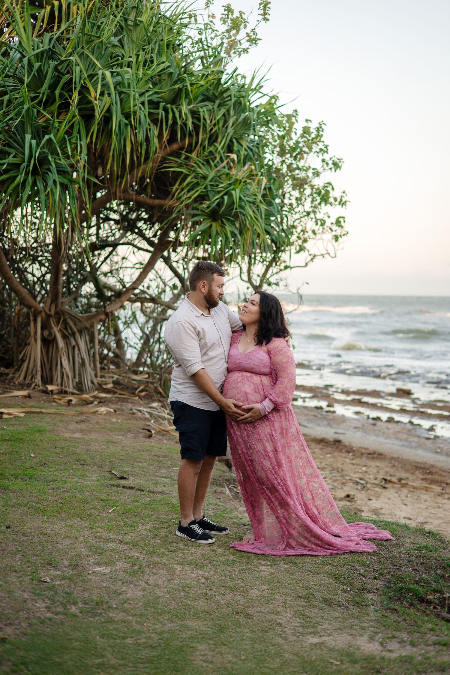 A couple shares a tender moment on a beach shoreline with lush tropical vegetation in the background at sunset.