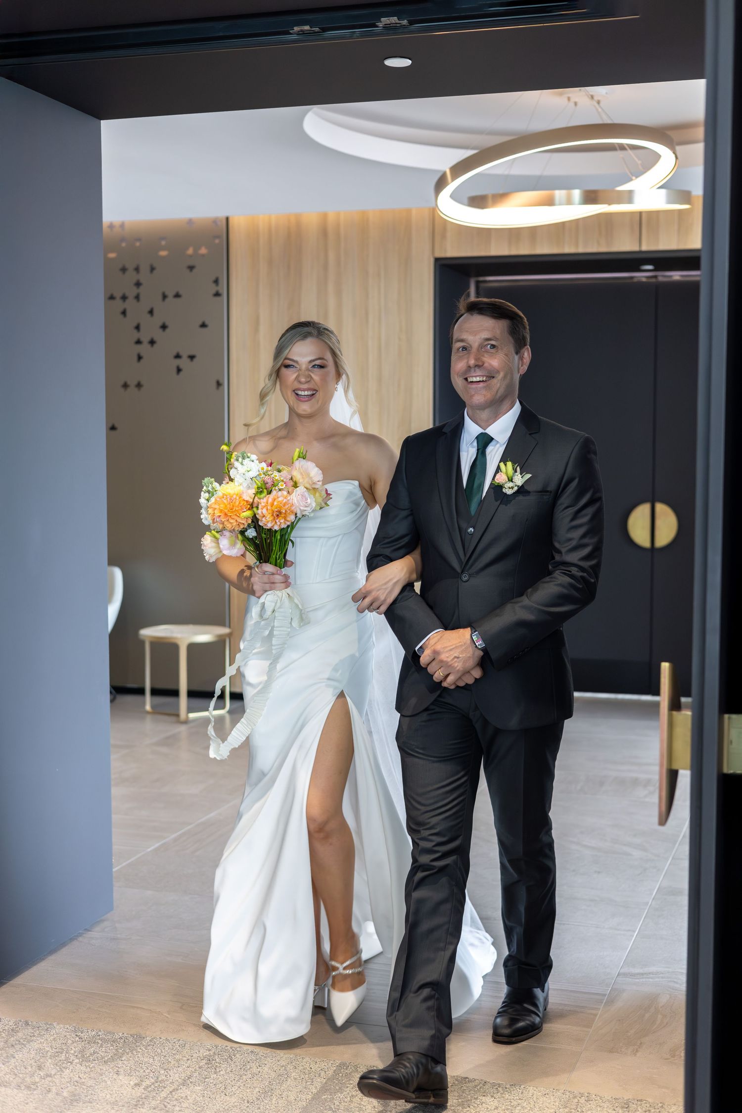 A couple in wedding attire shares joyful moments in a modern indoor venue with sleek grey walls and tiled floors.