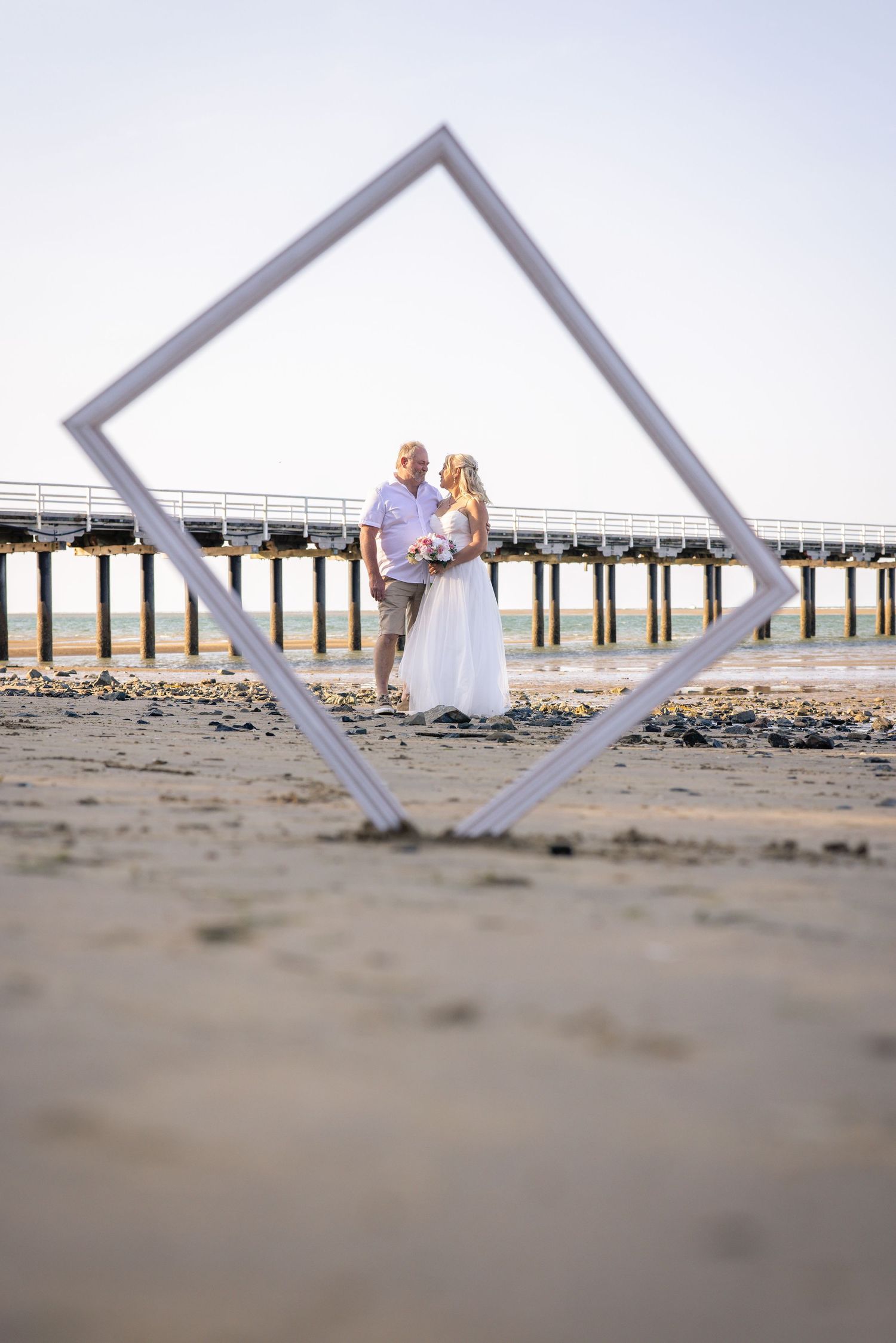 Wedding party photographed through geometric frames on a sandy beach with a pier in the background.