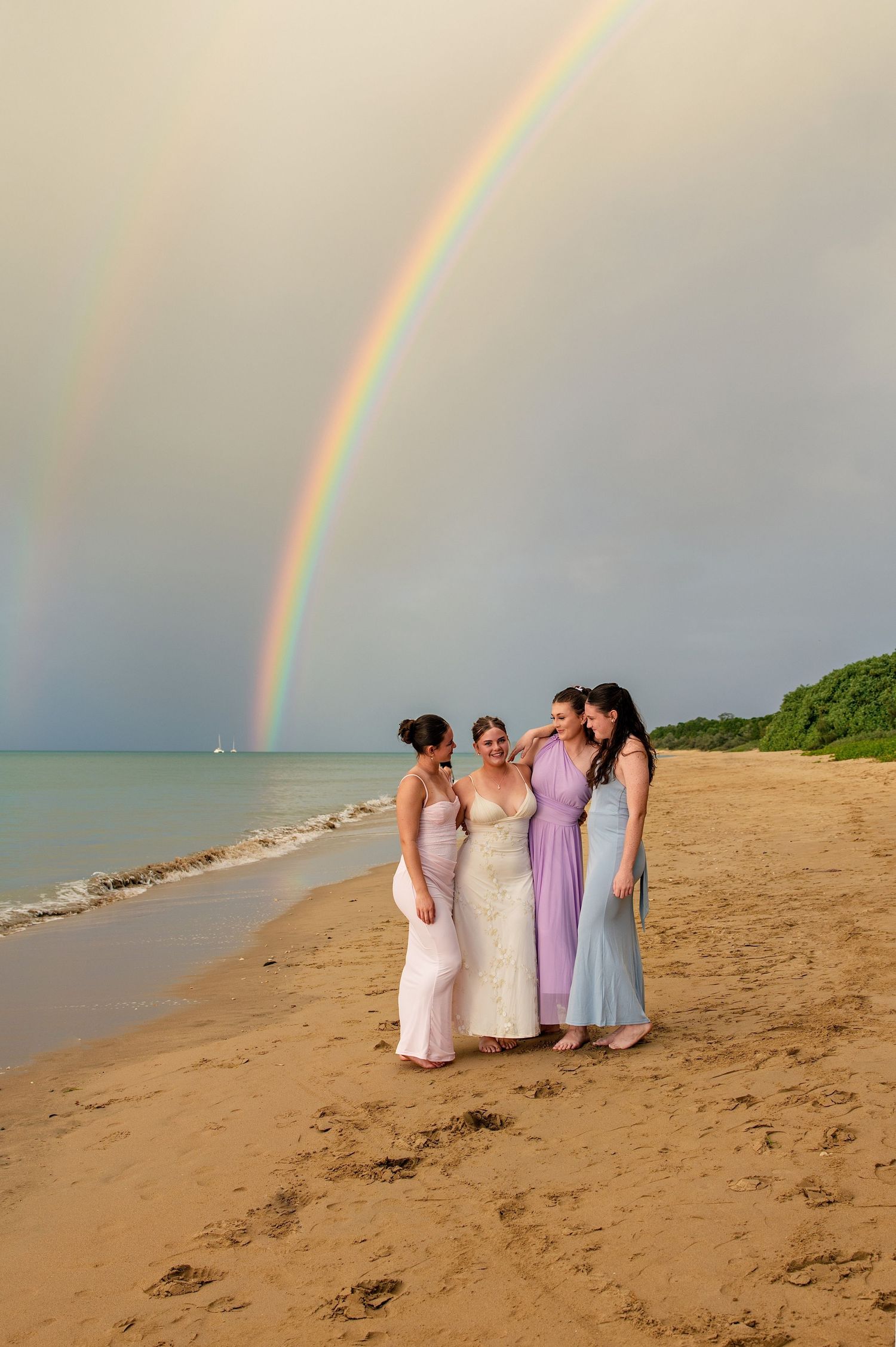 Bride and bridesmaids in pastel gowns pose on a sandy beach beneath a stunning double rainbow.