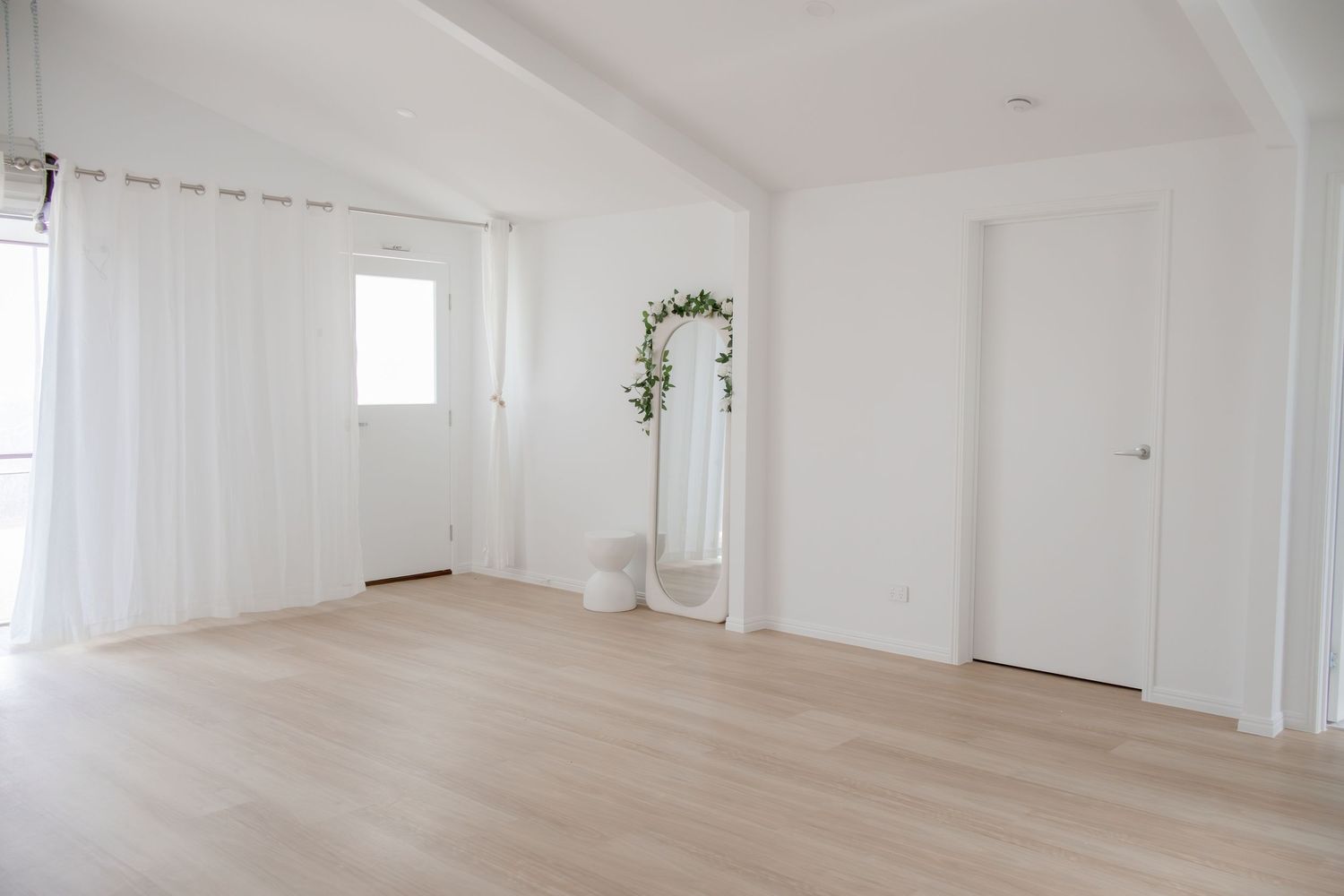 Bright minimalist white room with light wood floors, sheer curtains, and a decorative green floral arch.