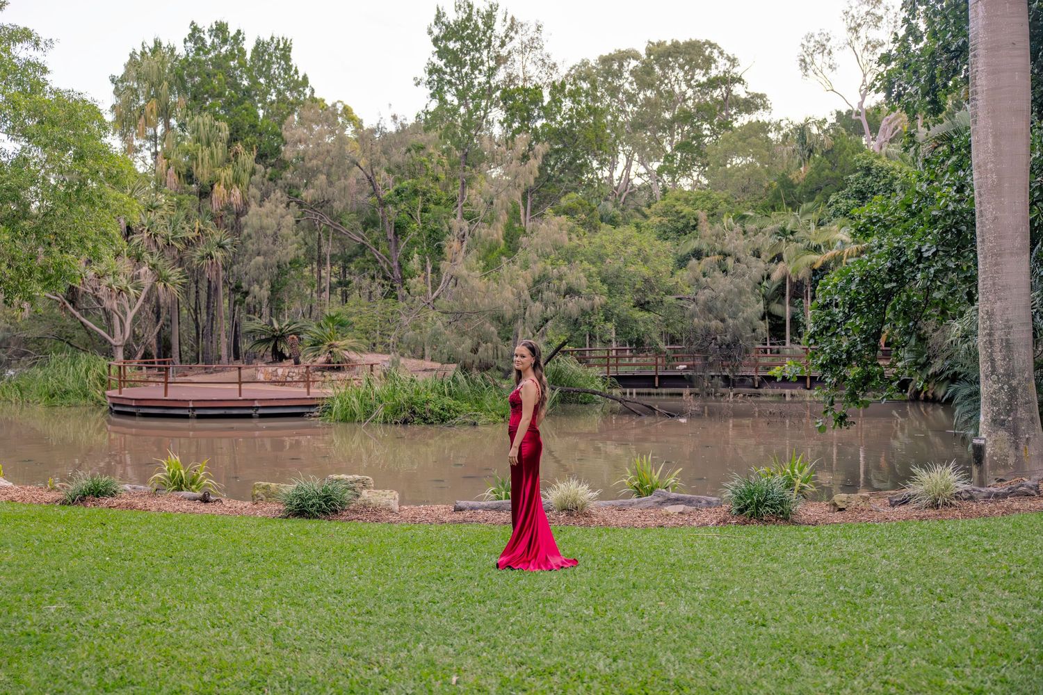 A figure in a flowing red gown stands gracefully on a manicured lawn near a tranquil pond surrounded by trees.