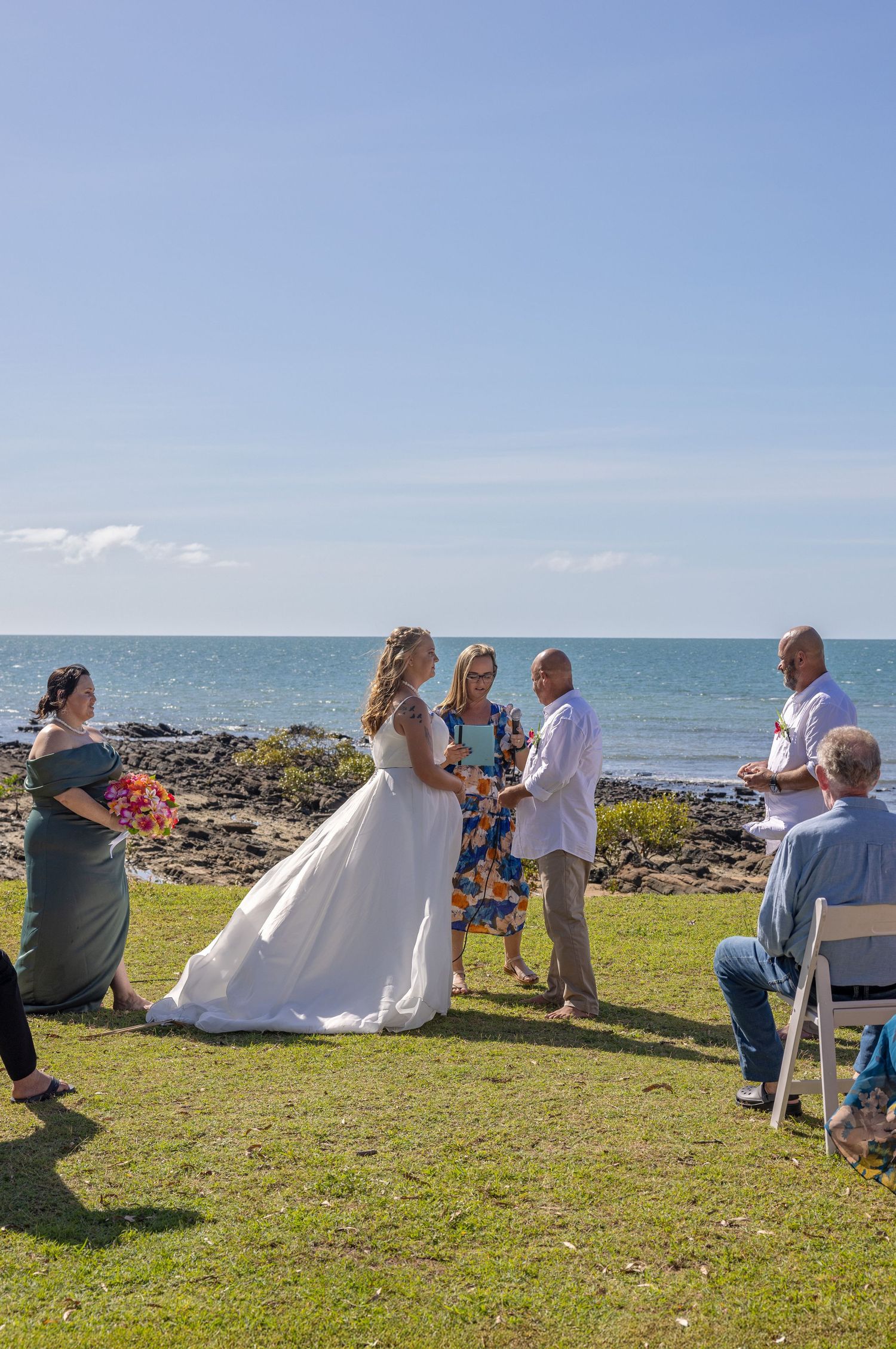 A small outdoor wedding ceremony takes place on a grassy cliff overlooking the ocean on a sunny day.