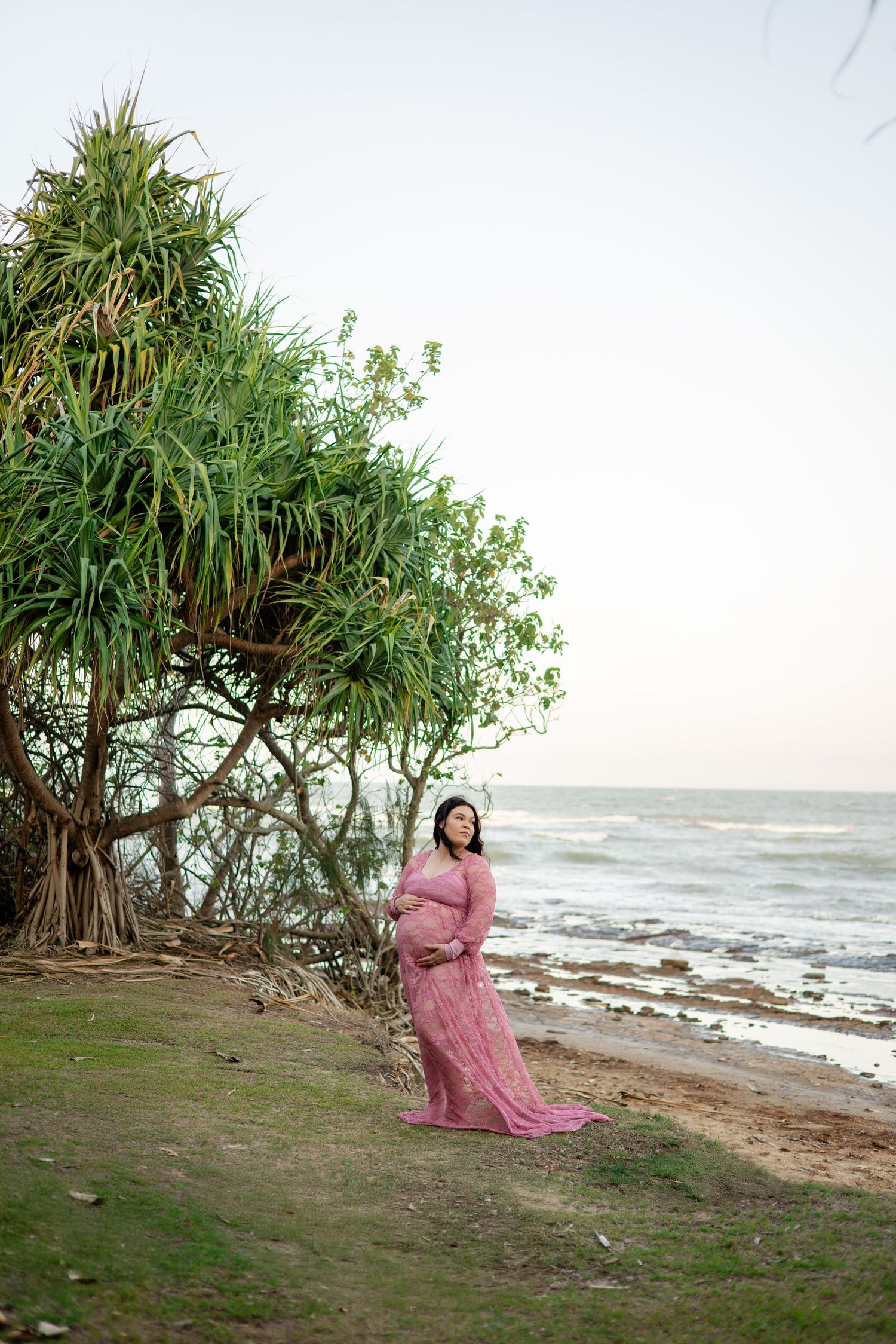 A flowing pink dress catches the breeze on a tropical beach with pandanus trees and ocean waves in the background.