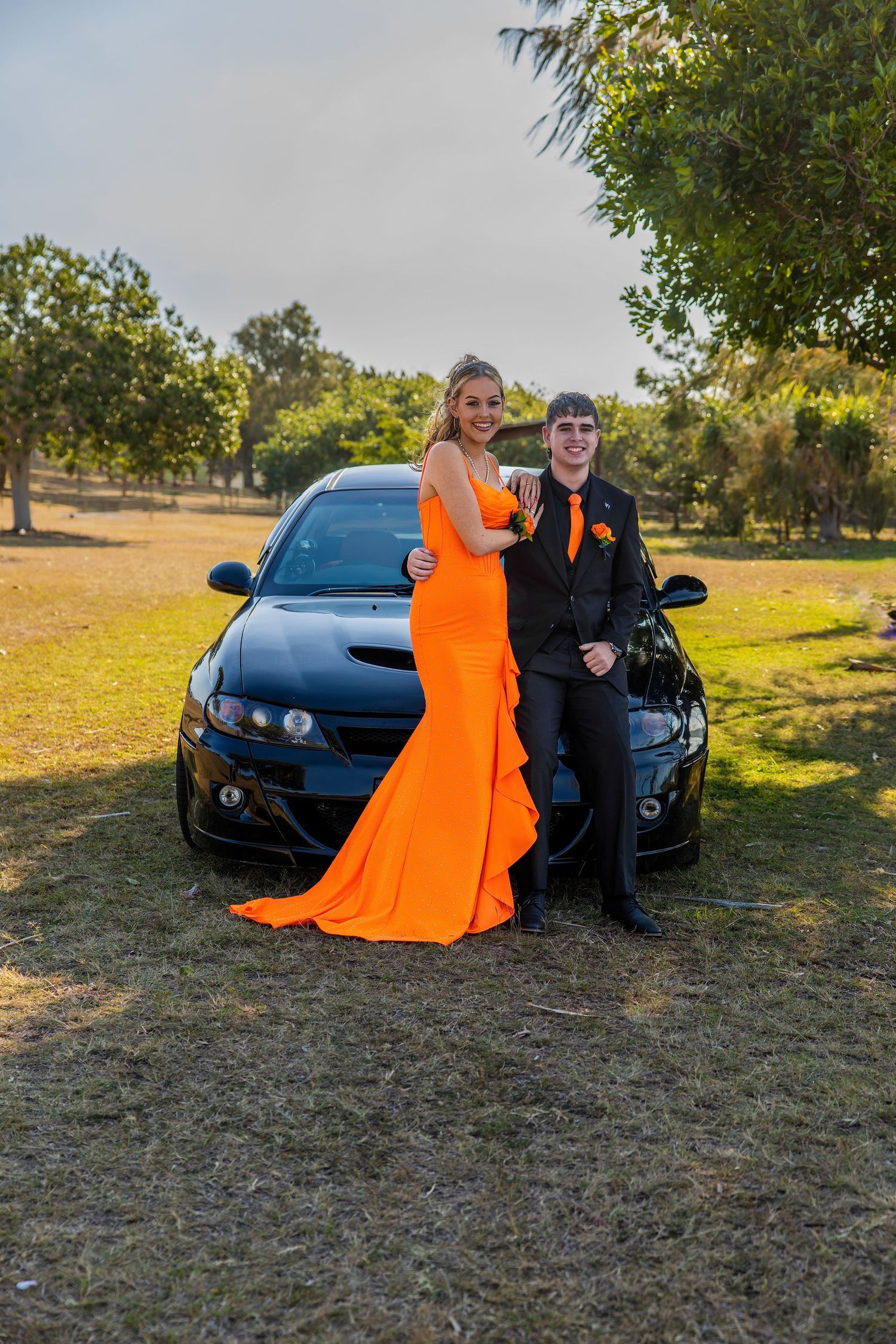 A couple in formal prom attire pose beside a black sports car, the girl wearing a bright orange gown.