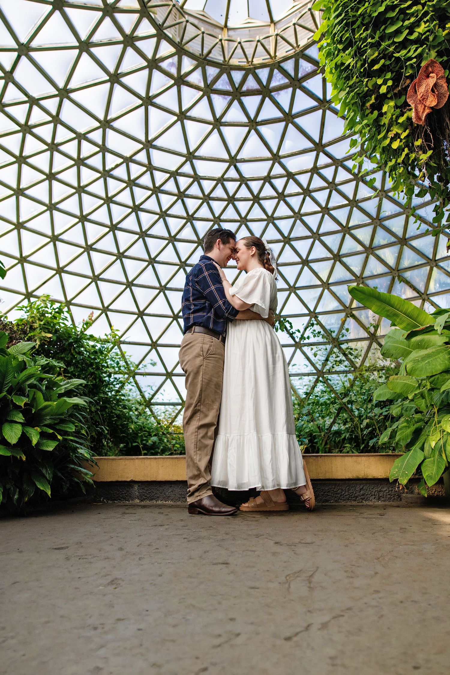 Couple embracing romantically inside a geodesic dome greenhouse surrounded by lush tropical plants.